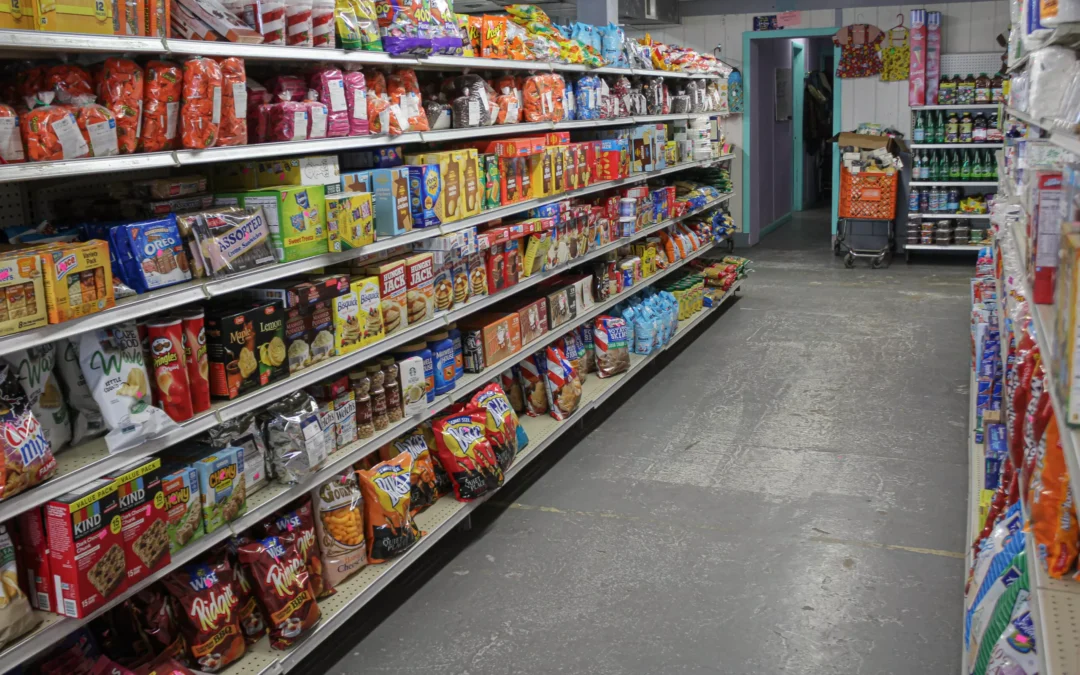 2.27.21-Leids-Market-Gallery-1-10-scaled-1 A grocery store aisle with shelves stocked with various packaged snacks, cereals, and household items; the floor is gray and the store appears well-lit.