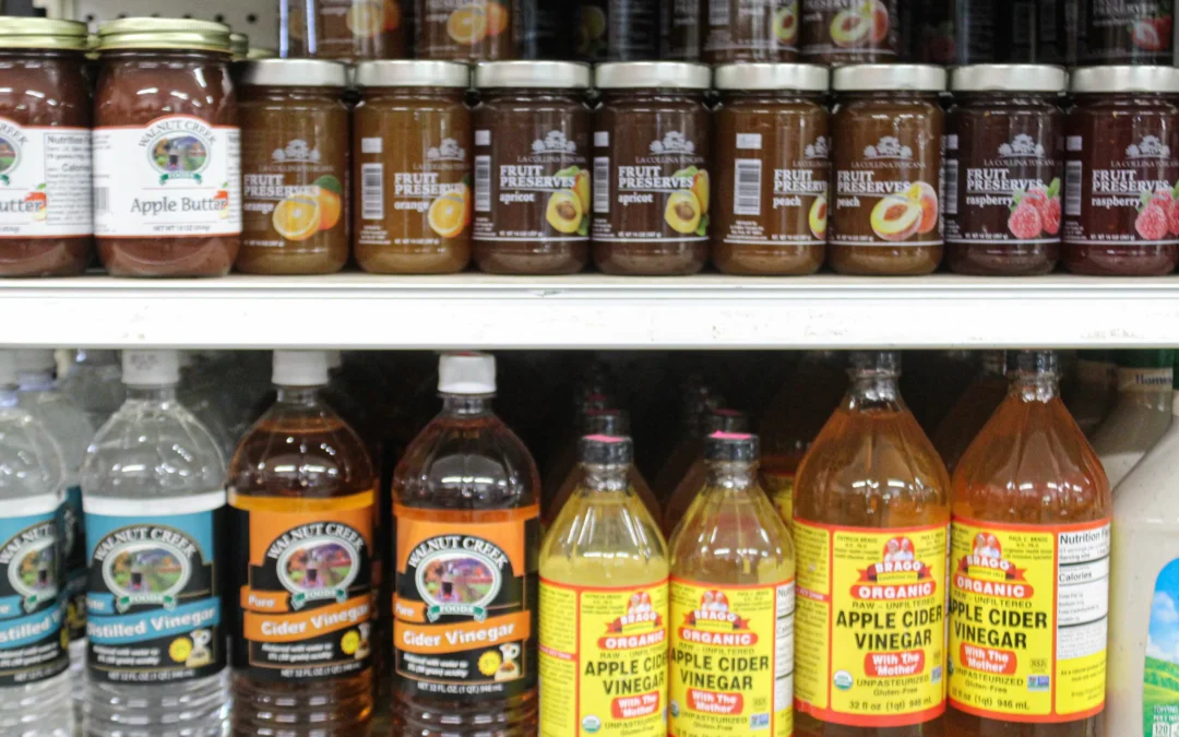 2.27.21-Leids-Market-Gallery-1-8-scaled-1 Jars of fruit preserves and apple butter are displayed on a shelf above bottles of distilled water and apple cider vinegar in a grocery store.