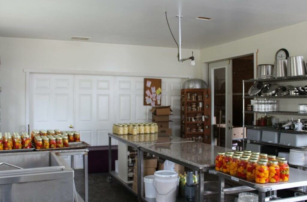 Homegrown Cannery Stand New Holland PA A kitchen with jars of preserved fruits arranged on stainless steel tables, shelves with kitchen supplies, a bulletin board, and a pantry in the background.