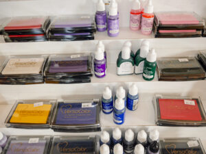 A display of various Versacolor ink pads and bottles of liquid ink in different colors, organized on a white shelf. Prices are marked on individual items.