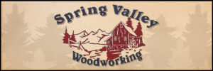 Logo for Spring Valley Woodworking, featuring a red barn, trees, and mountains in the background, with the company name in curved text above and below the image.