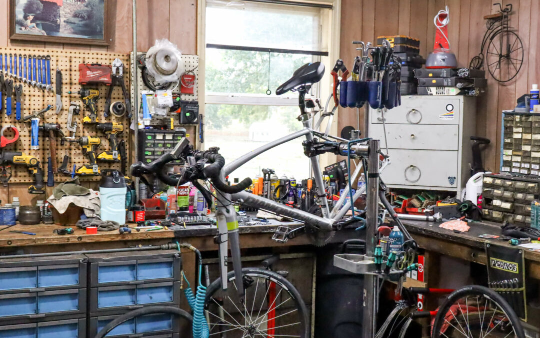 Weaver's Bike Shop Ephrata PA A cluttered workshop with a bicycle on a repair stand surrounded by various tools, drawers, and equipment. A window in the background reveals a view of greenery outside.