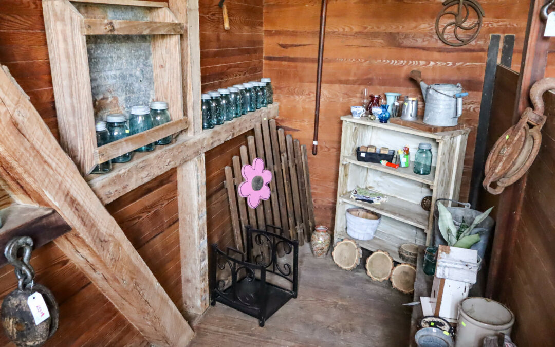 9.28.24 Pine Hill Farms Antique Gallery 1 A rustic wooden antique store with shelves holding jars, bottles, and miscellaneous items. A folding chair with a pink flower is against the wall, and a wooden display of small logs is on the floor.