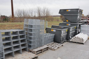 Outdoor storage area with stacks of metal grates, gray industrial hoppers, and bundles of white pipes on wooden pallets. Background shows bare trees and a grassy field under a cloudy sky.