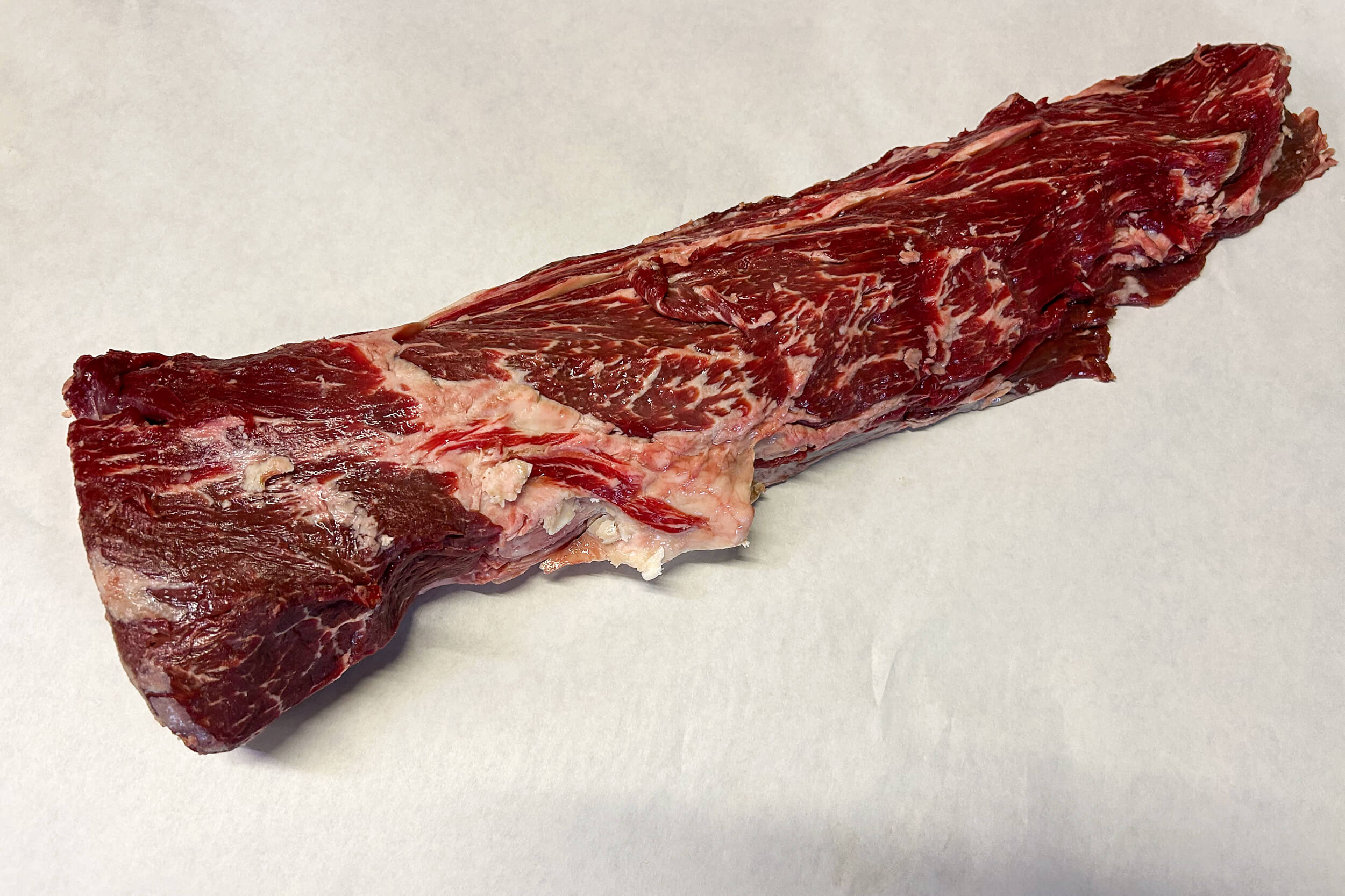 A raw, untrimmed piece of beef skirt steak resting on a white surface.