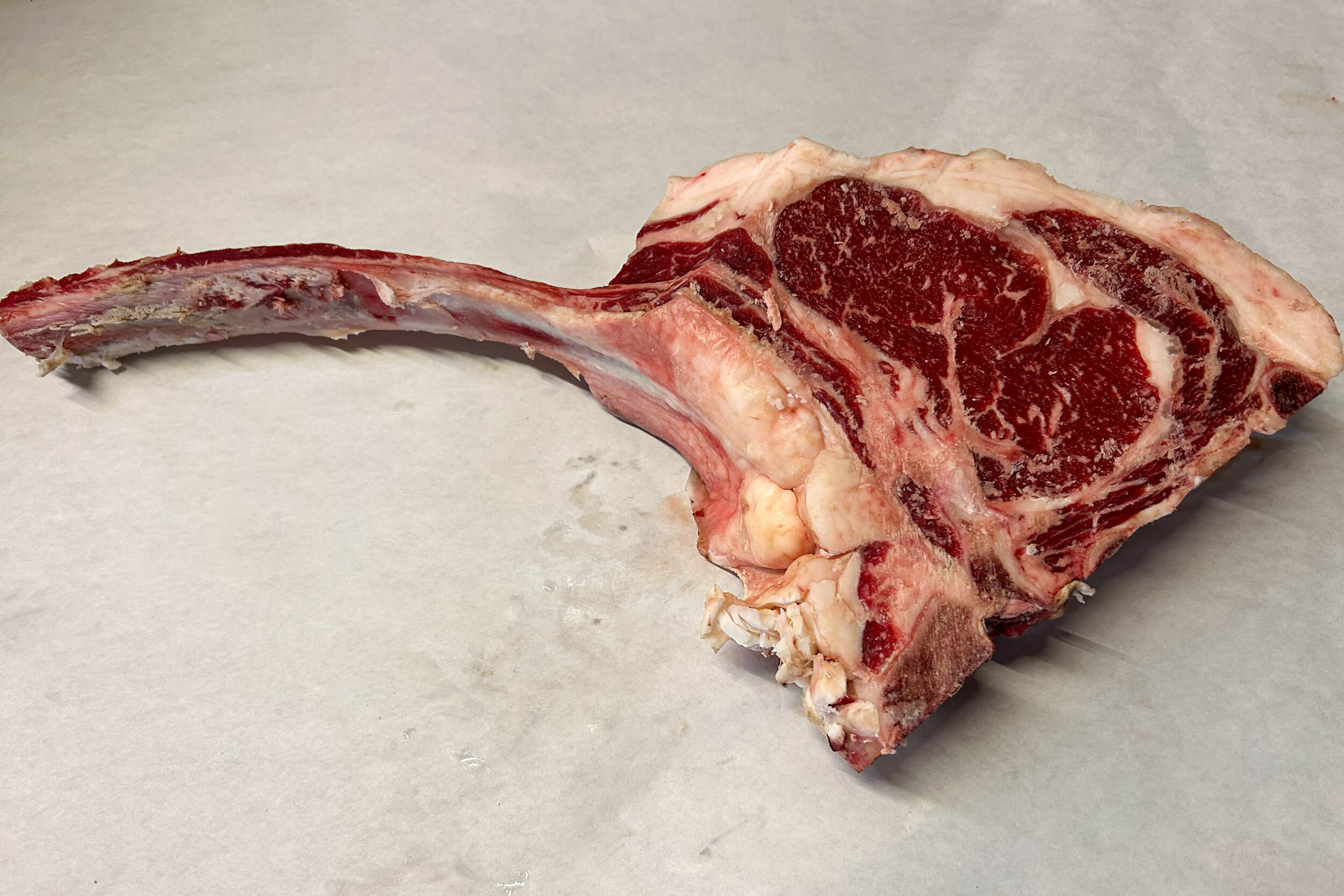 A raw tomahawk ribeye steak with a long bone is placed on a light-colored surface.