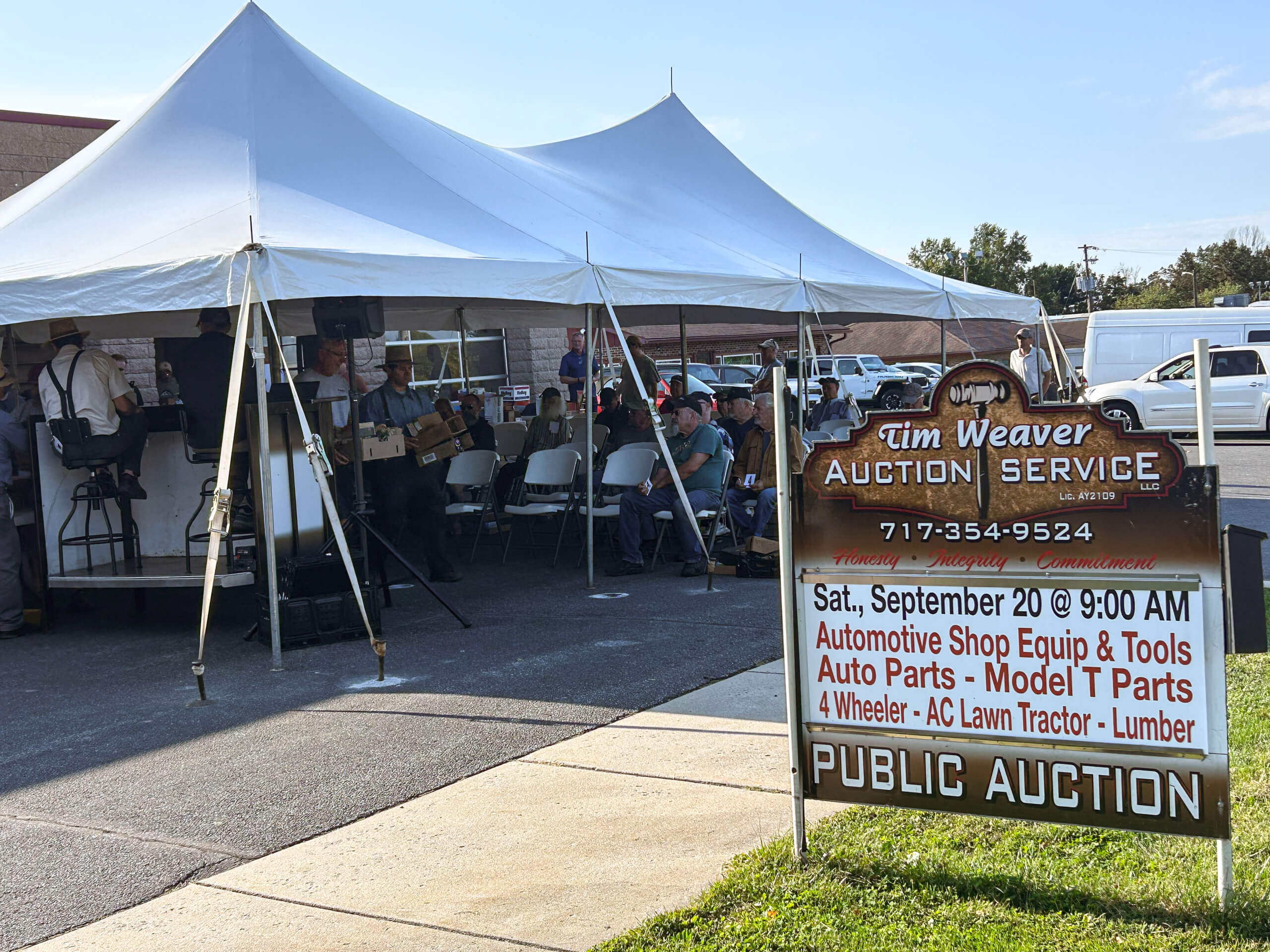 Tim Weaver Auction Service Lancaster County PA People sit under a white canopy tent at a public auction event; a sign in the foreground advertises auction details, including date, time, and items for sale.
