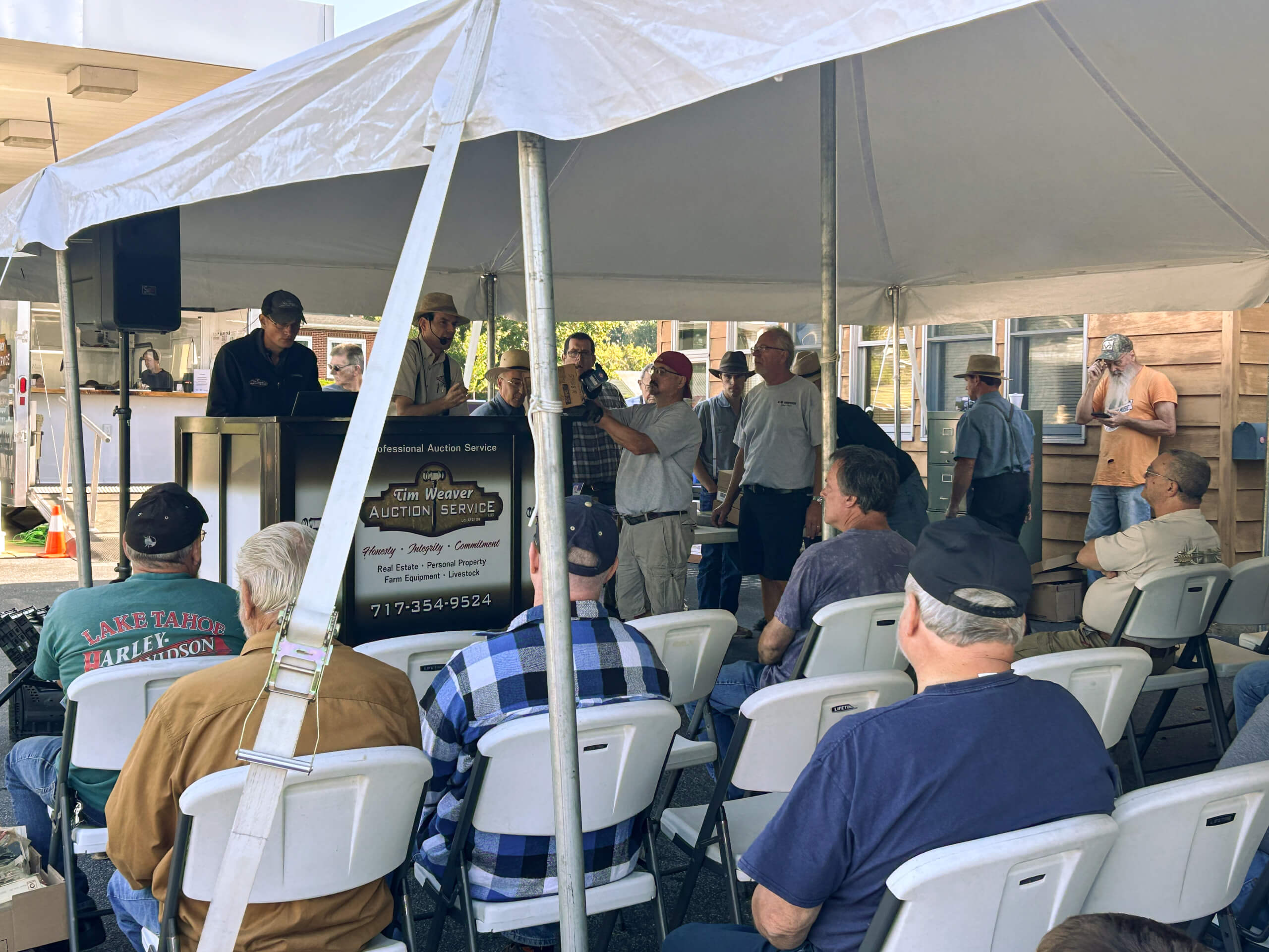 Tim Weaver Auction Service Lancaster County PA A group of people sit under a white tent, watching an auctioneer at a podium as others stand and observe the event.