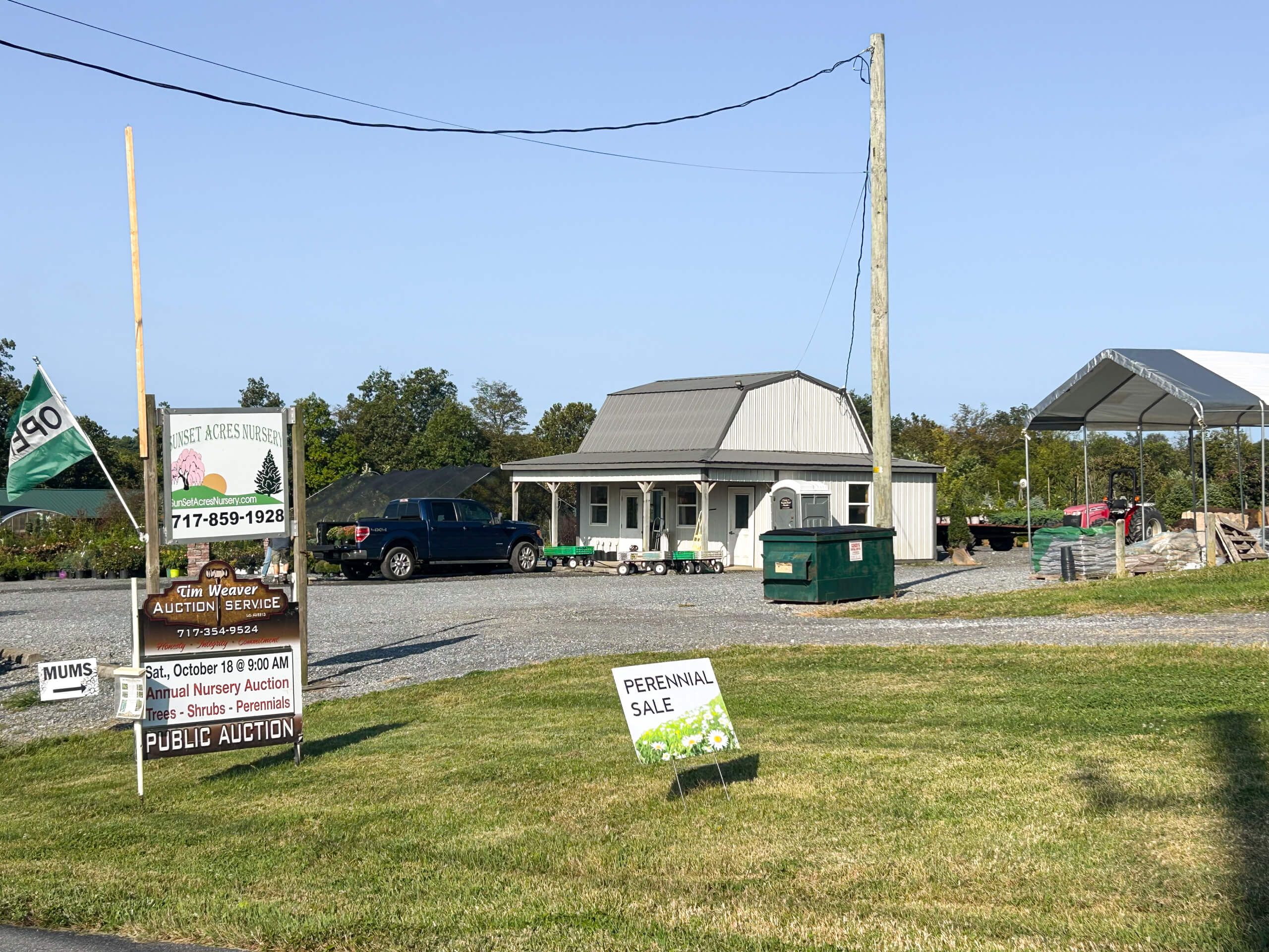 Tim Weaver Auction Service Lancaster County PA Small building with a metal roof, a black SUV parked outside, signs advertising a public auction and perennial sale, and a green dumpster nearby on a gravel lot.