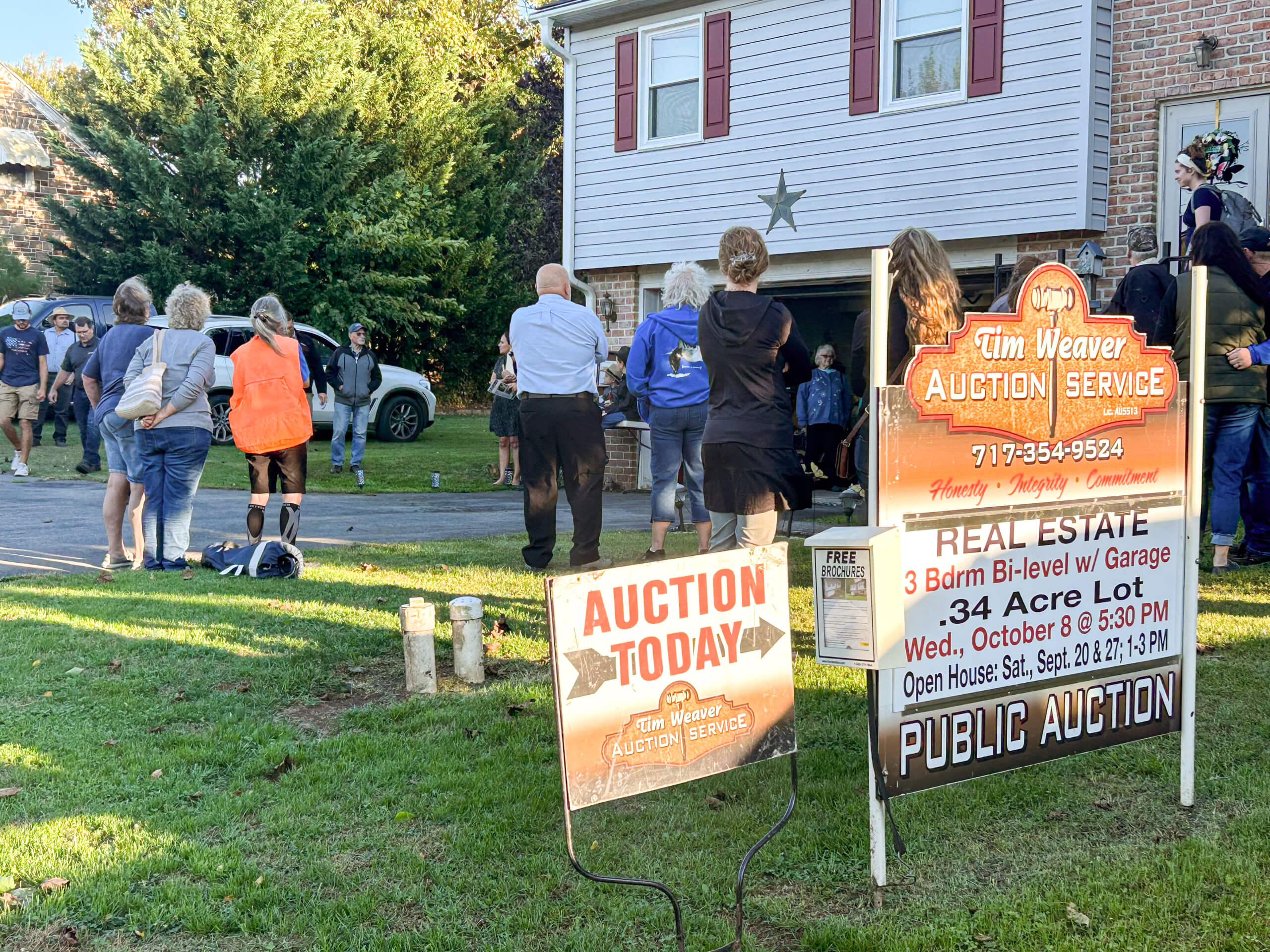 Tim Weaver Auction Service Lancaster County PA A group of people gathers outside a two-story house for a public real estate auction, with auction signs displayed on the front lawn.