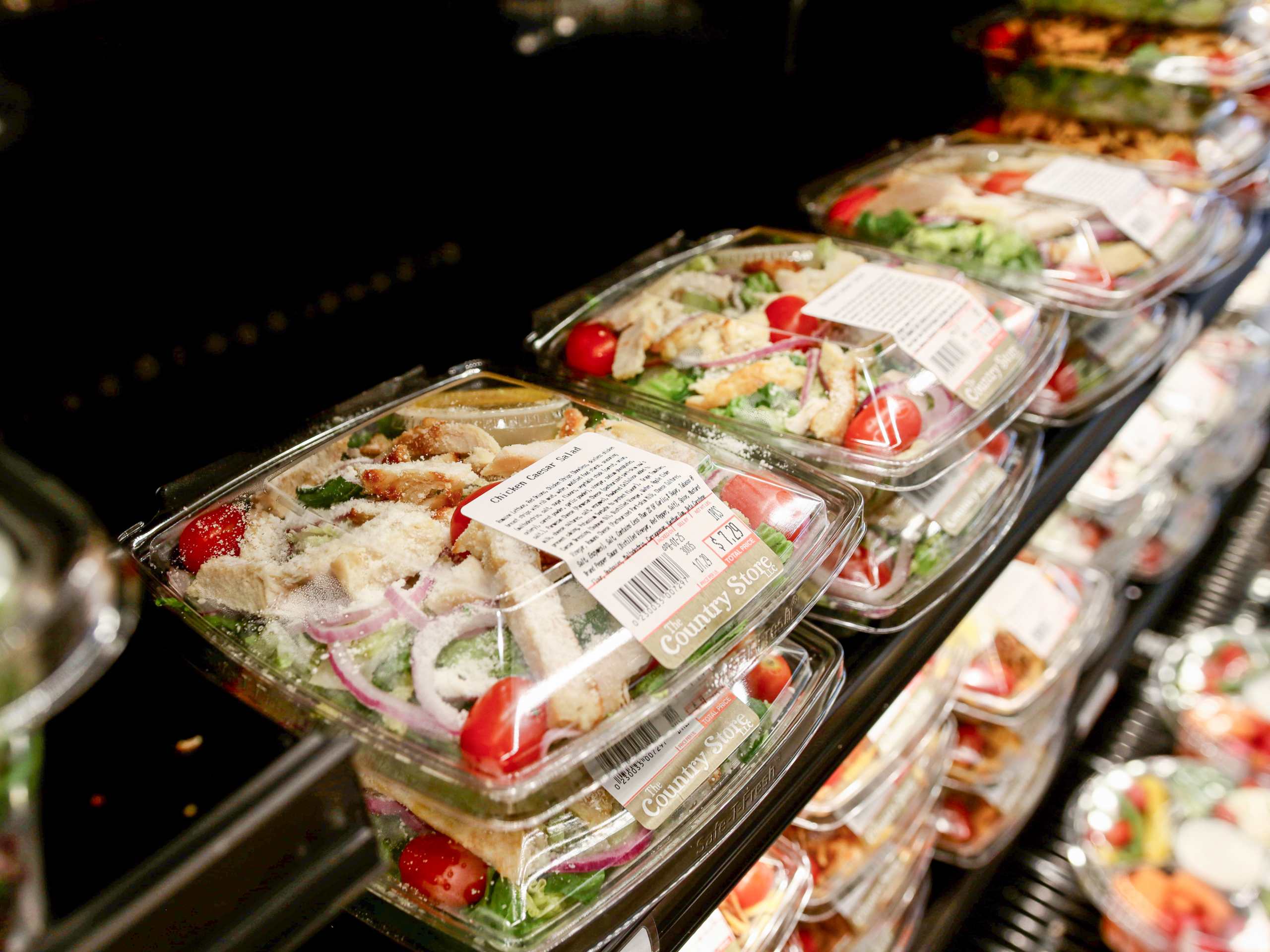 The Country Store Mount Joy PA Plastic containers of prepared salads with labels stacked on a refrigerated grocery store shelf.