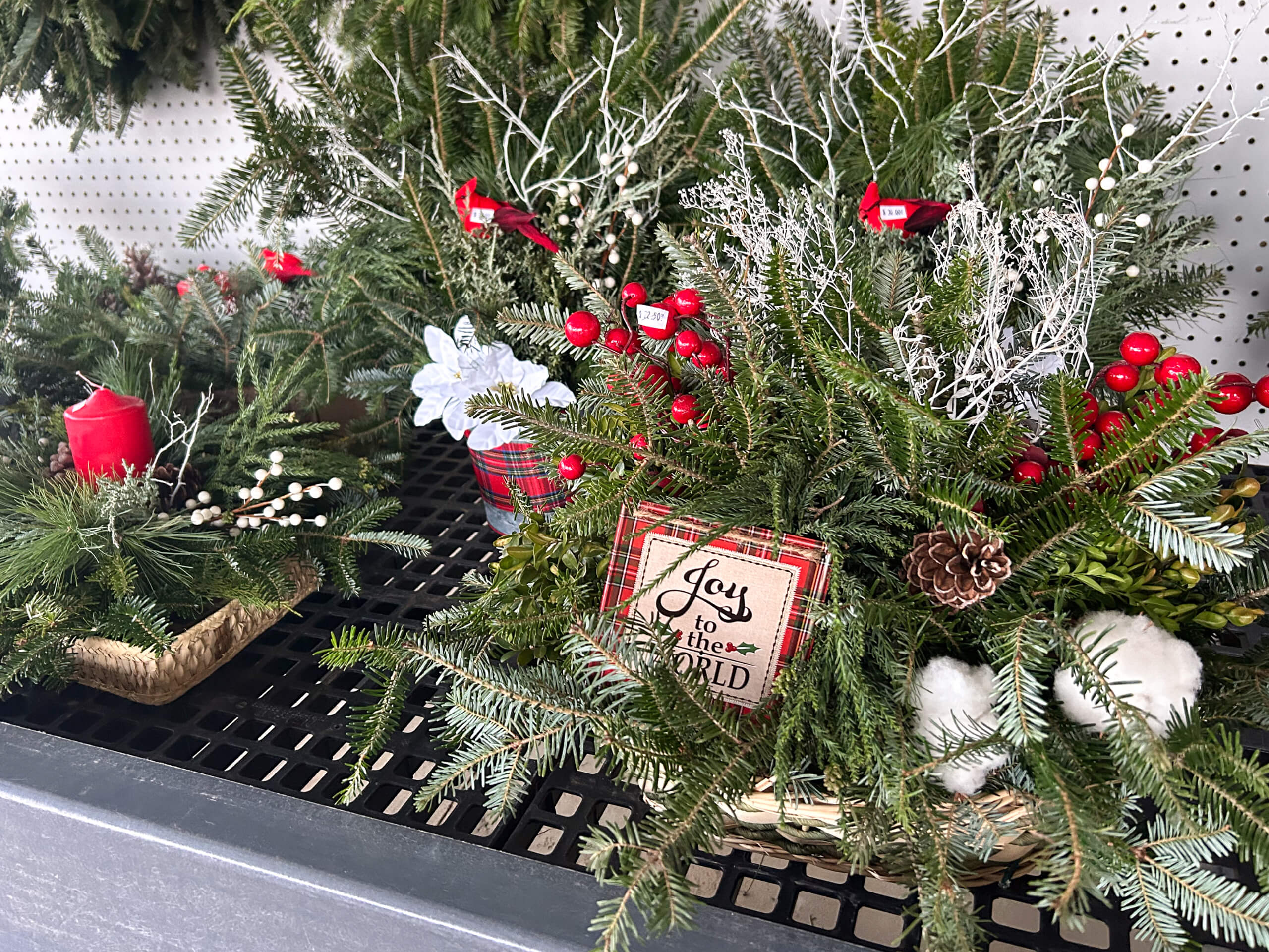 Stoltzfus Produce & Marketfare New Holland PA Holiday-themed floral arrangements with pine branches, red berries, pinecones, candles, and a sign reading "Joy to the World" displayed on a black metal shelf.