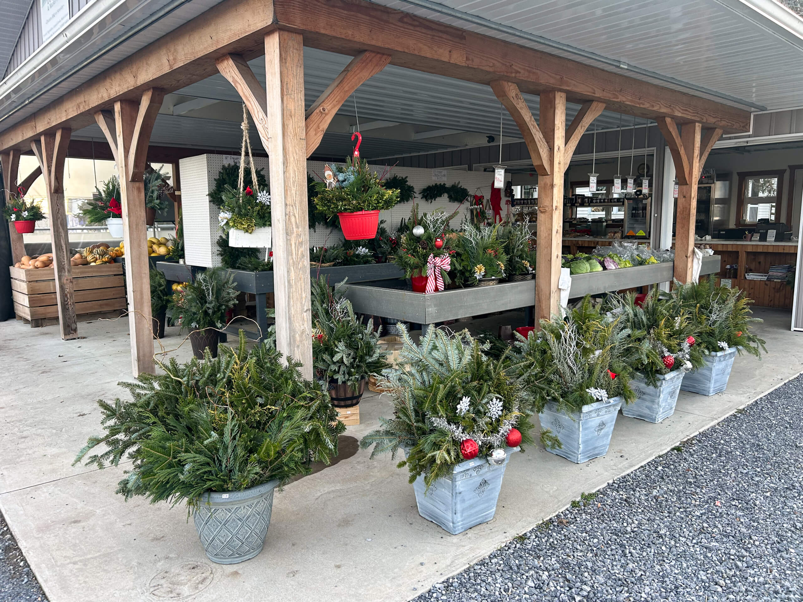 Stoltzfus Produce & Marketfare New Holland PA Outdoor market stand with wooden beams displaying various potted evergreen arrangements, hanging baskets, and produce crates under a covered area.
