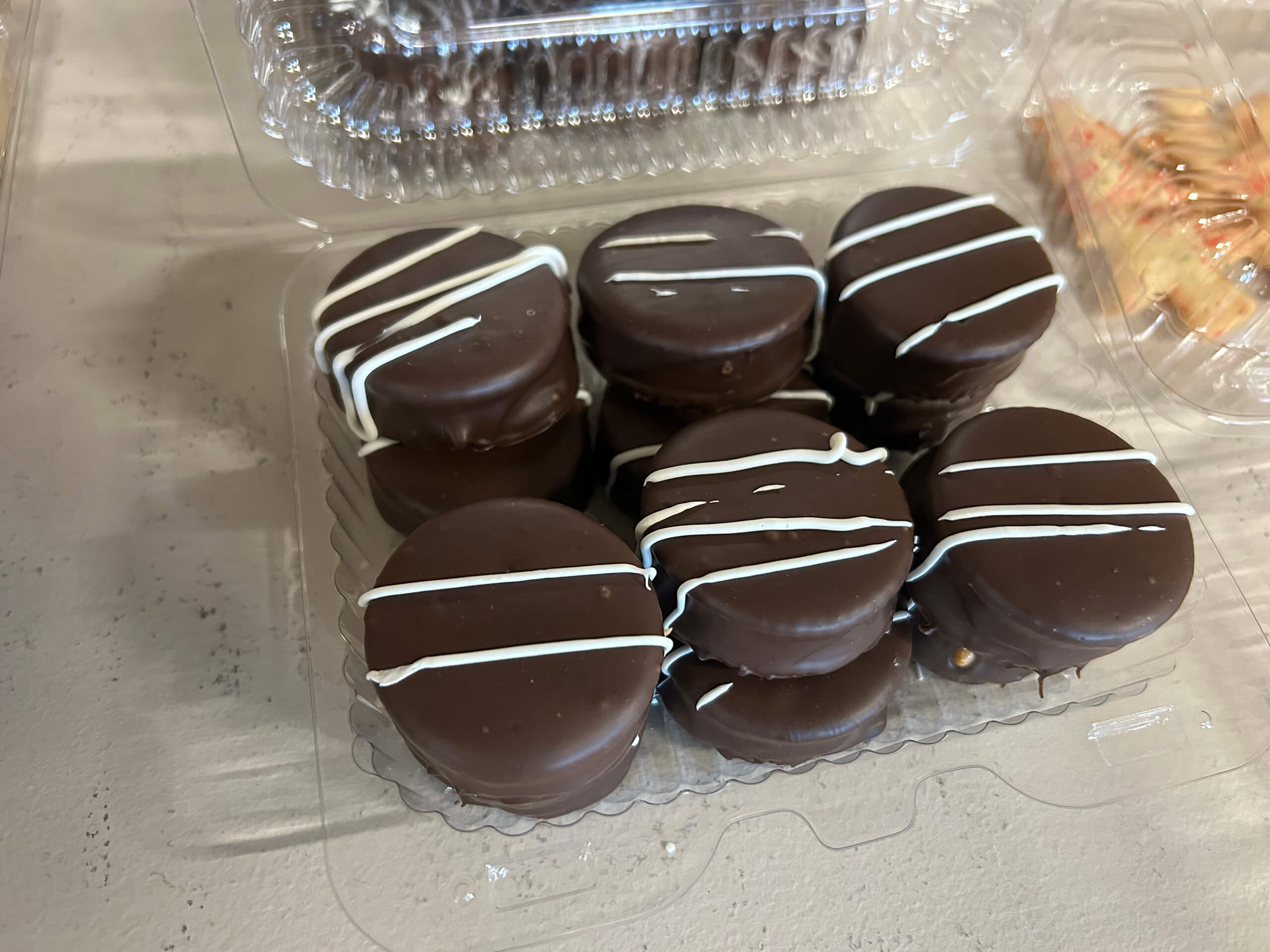 Turtle Hill Farm Market Leola PA Six chocolate-covered sandwich cookies with white chocolate drizzle are arranged in a clear plastic container on a light-colored surface.