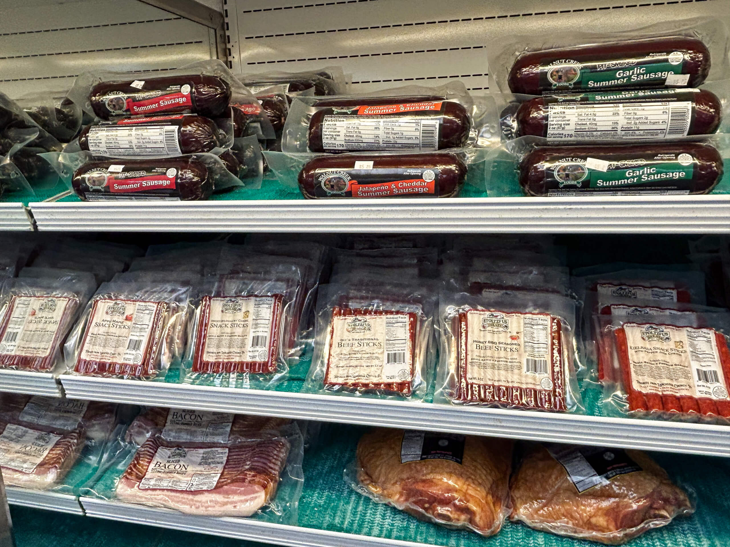 Sensenig Poultry Lititz PA Packages of sausages, meat sticks, bacon, and other processed meats are displayed on metal shelves in a refrigerated grocery store section.