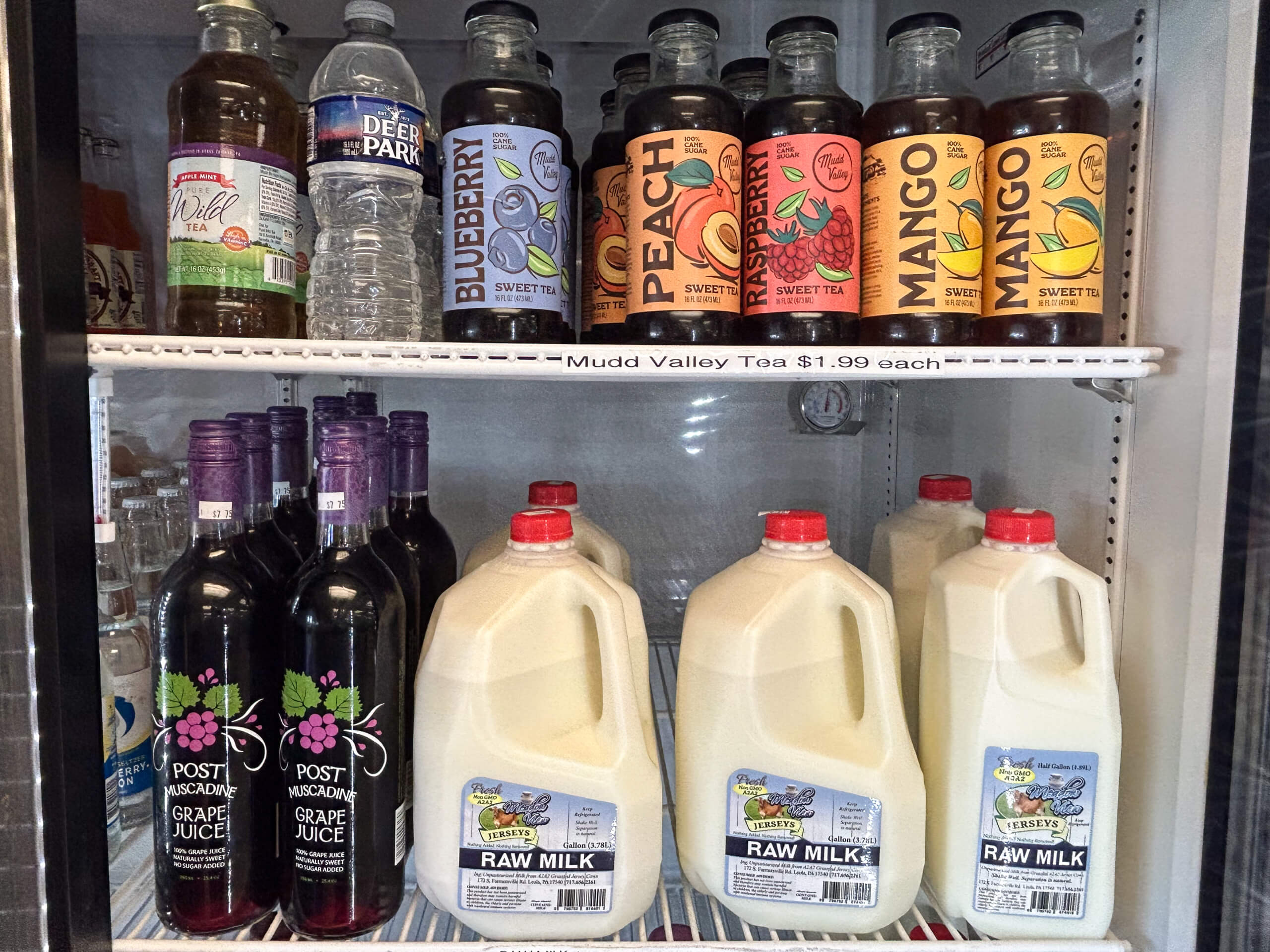 Sensenig Poultry Lititz PA Refrigerator shelves with bottled water, flavored teas, grape juice, and gallon jugs of raw milk. A label shows "Mudd Valley Tea $1.99 each.