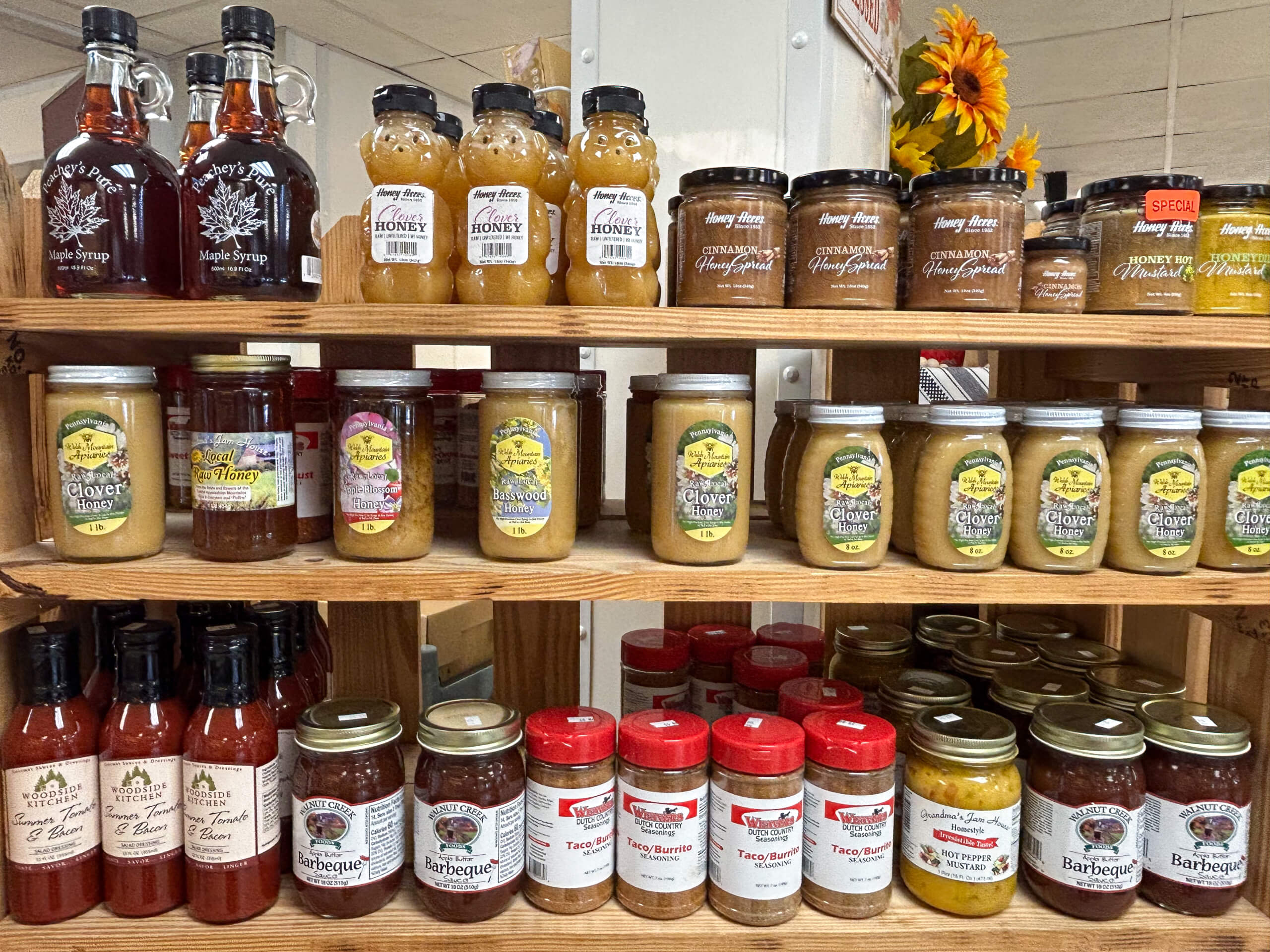 Sensenig Poultry Lititz PA Wooden shelves displaying jars and bottles of maple syrup, honey, jams, sauces, and seasonings arranged in rows at a store.