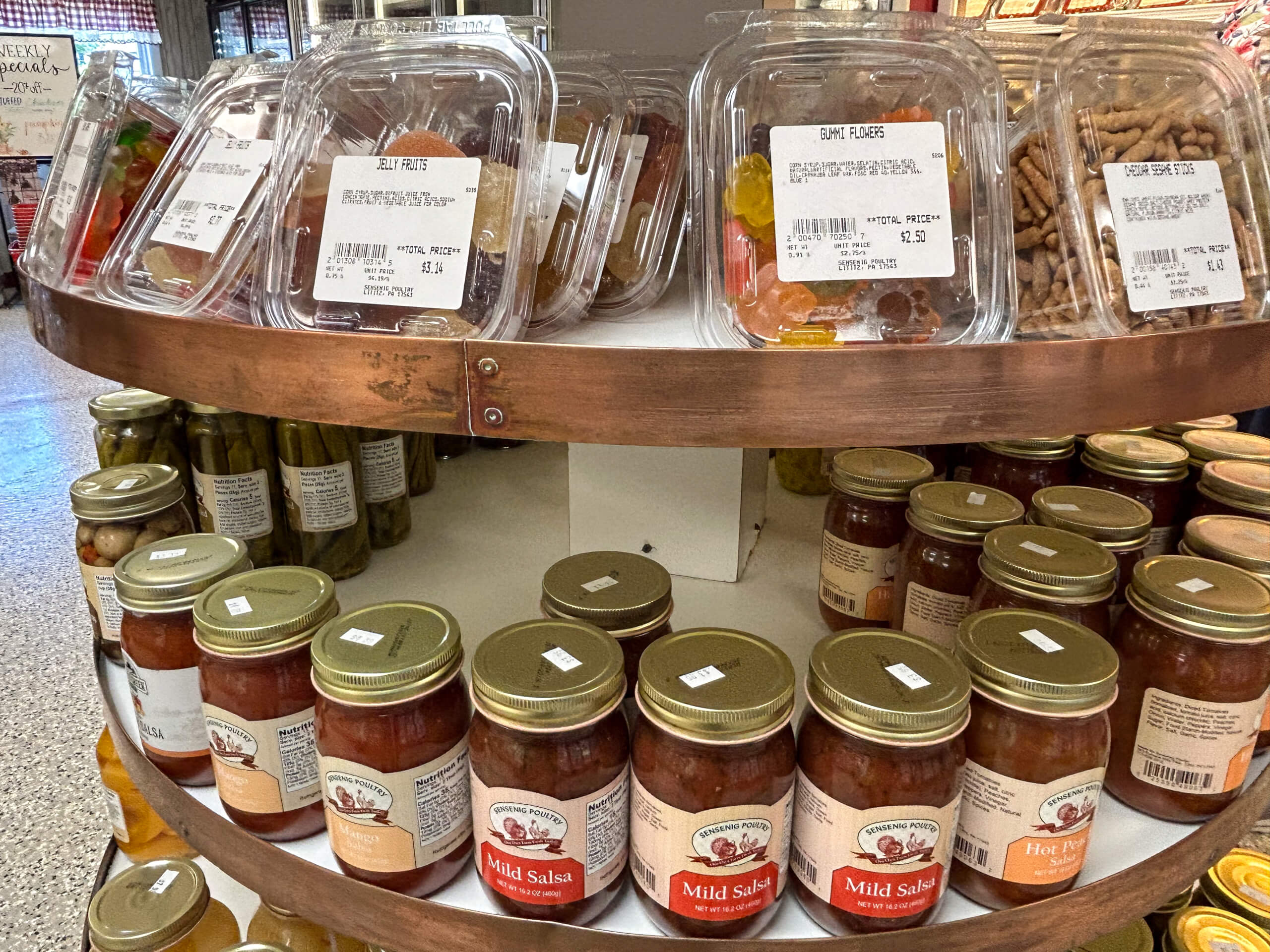 Sensenig Poultry Lititz PA Shelf displaying containers of dried fruit and candy on the upper tier, with jars of salsa and pickles on the lower tier in a grocery store.