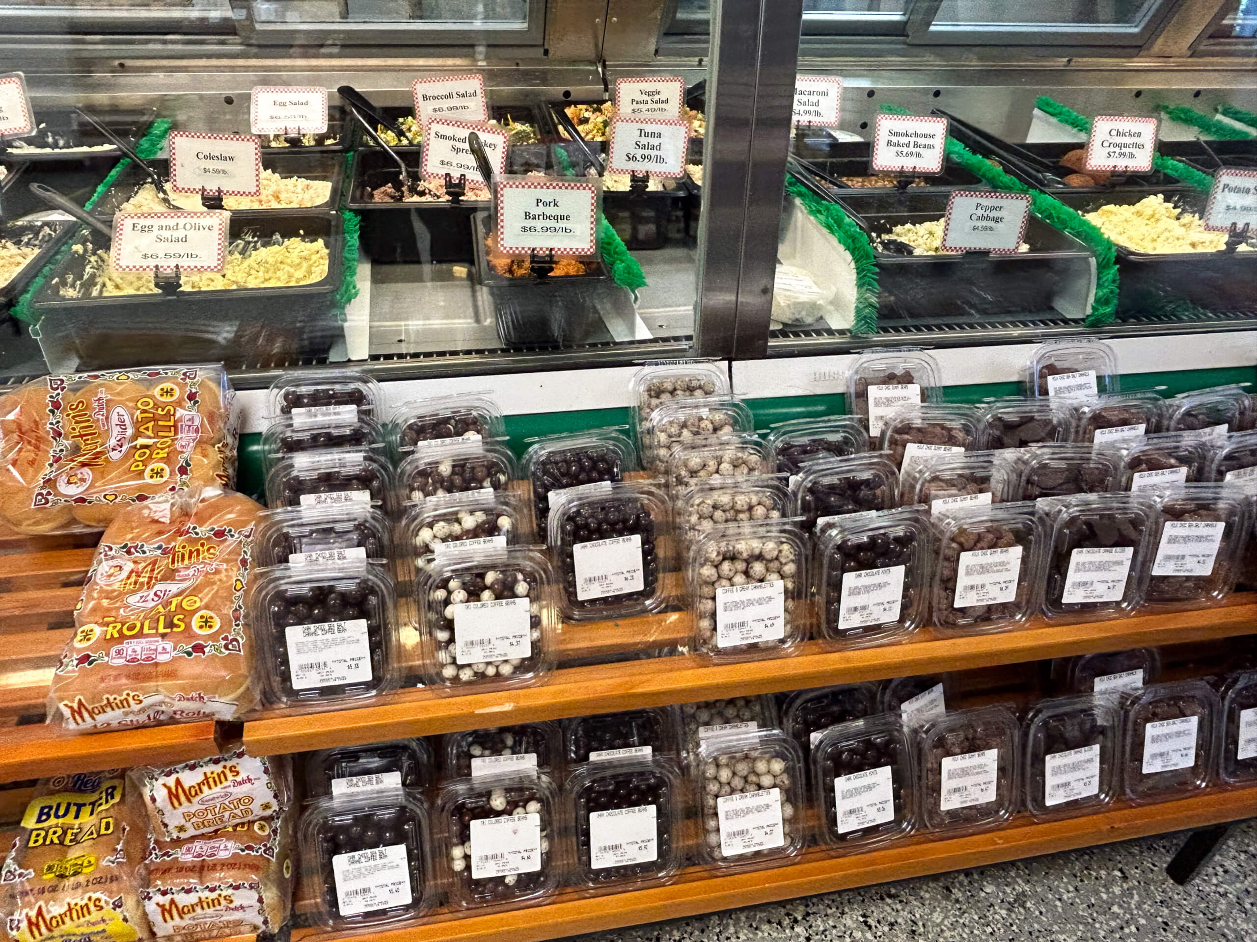 Sensenig Poultry Lititz PA Prepared food deli counter with various salads in trays behind glass, and several shelves stocked with packaged baked goods and containers of chocolate-covered nuts or candies in front.
