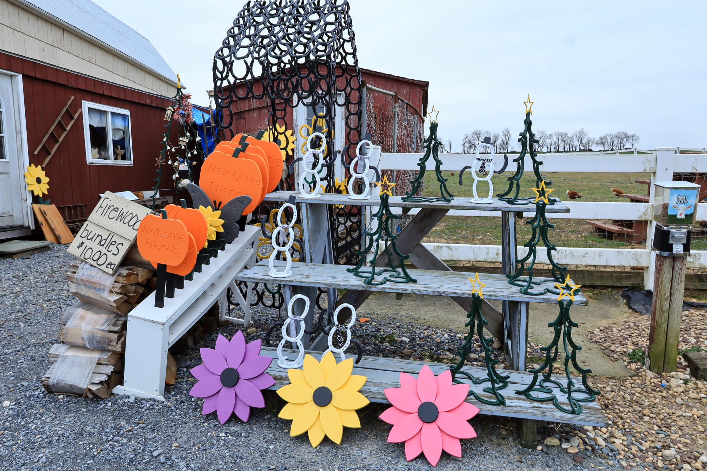 Busy Bee's Farm Market Ronks PA Outdoor display of metal and wooden decorative items for sale, including pumpkins, flowers, snowmen, and Christmas trees, arranged on shelves near a barn.