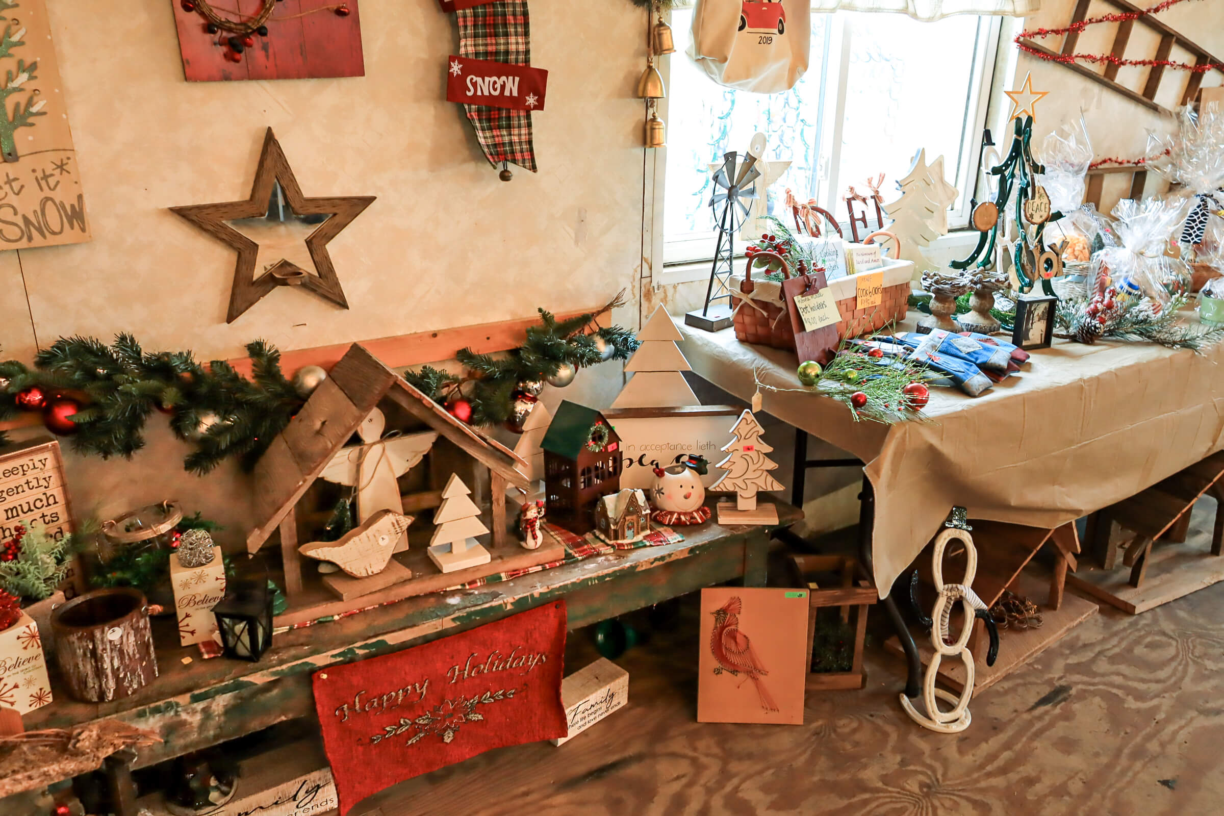 Busy Bee's Farm Market Ronks PA A display of assorted Christmas decorations including signs, ornaments, garlands, and gift baskets arranged on a bench and table by a window.