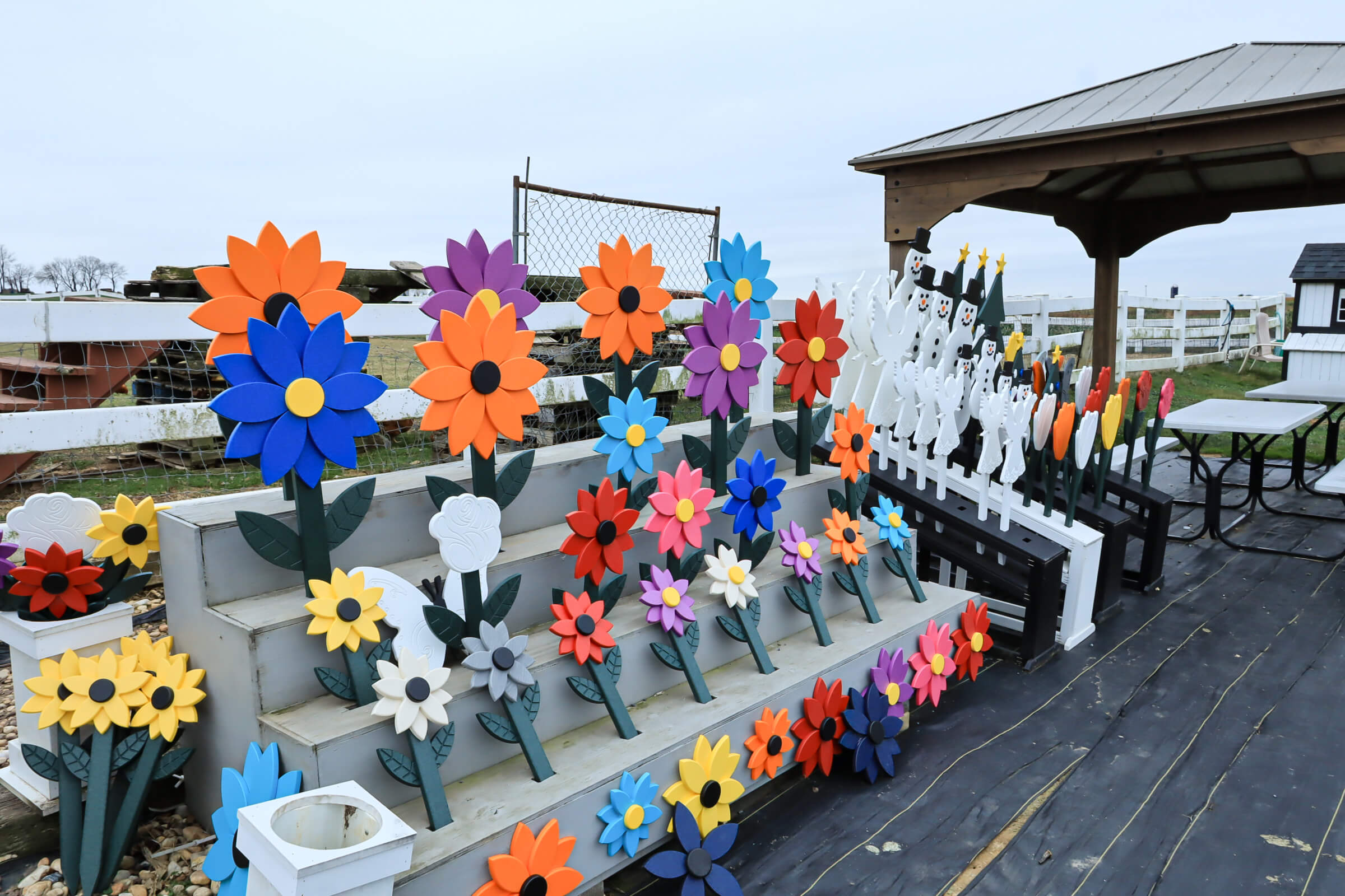 Busy Bee's Farm Market Ronks PA A display of colorful wooden flower decorations is arranged on tiered shelves and fenced panels outdoors near a gazebo.