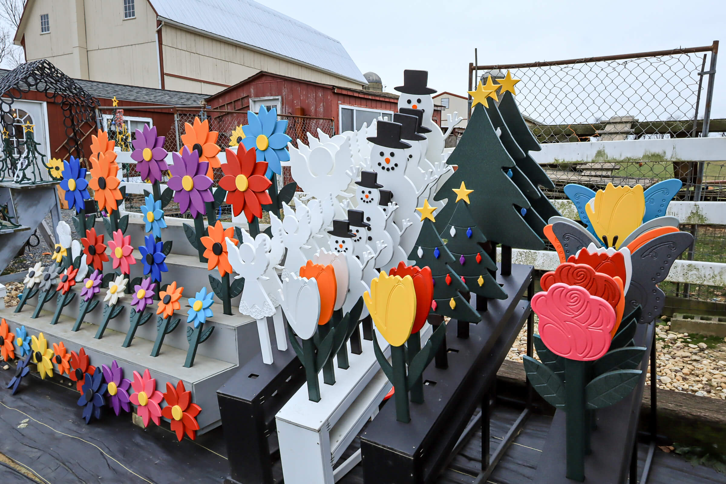 Busy Bee's Farm Market Ronks PA Rows of colorful wooden lawn ornaments, including flowers, tulips, snowmen, and Christmas trees, displayed outdoors near a barn and a chain-link fence.