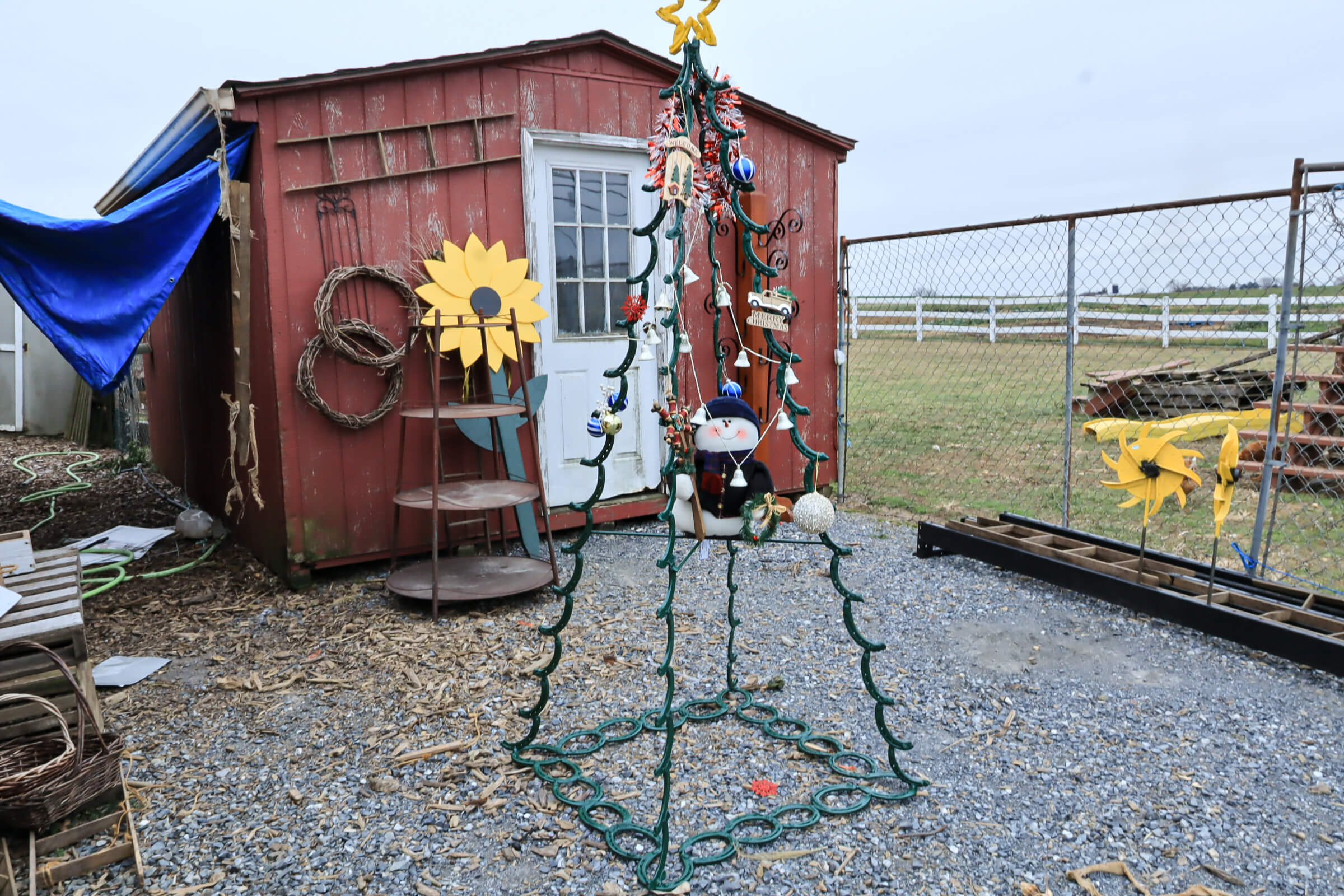 Busy Bee's Farm Market Ronks PA A metal frame shaped like a Christmas tree, decorated with ornaments and a snowman, stands outside a red shed on a gravel surface near a fenced yard.