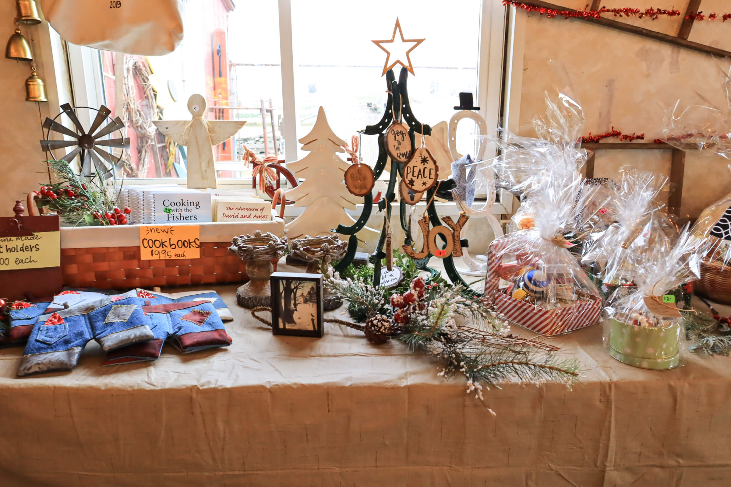 Busy Bee's Farm Market Ronks PA A table displays handmade crafts, baked goods, cookbooks, holiday decorations, and gift baskets in front of a window with festive ornaments and garland.