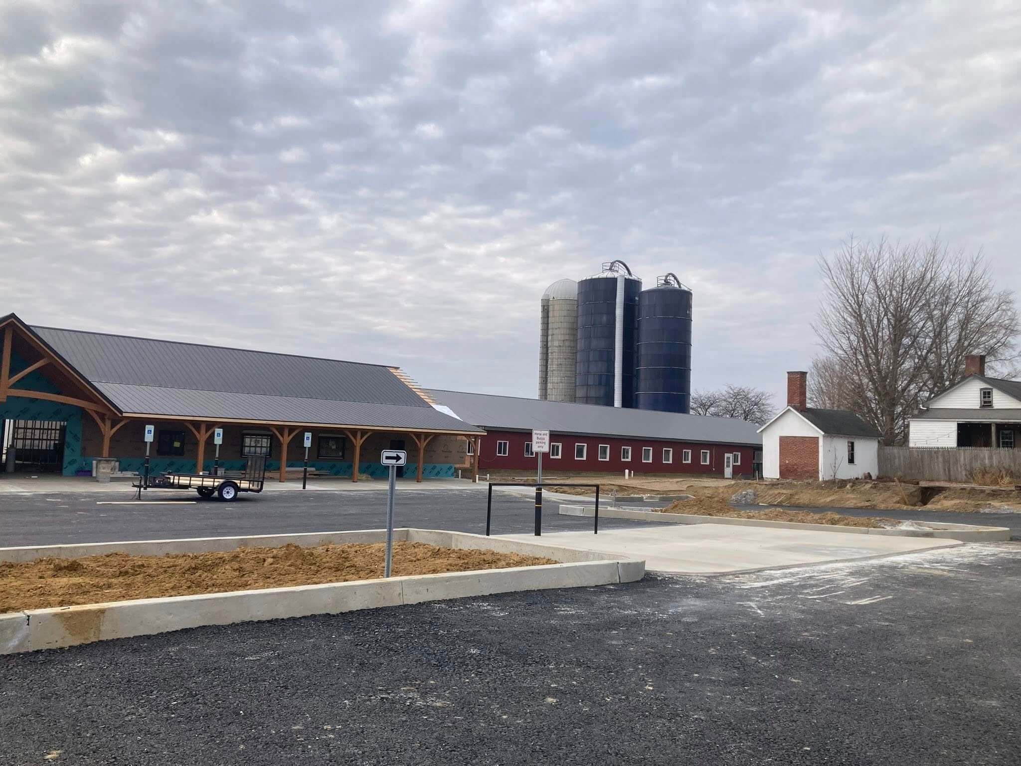 A farm scene with a long red barn, three tall silos, a small white house, and a parking lot under a cloudy sky.