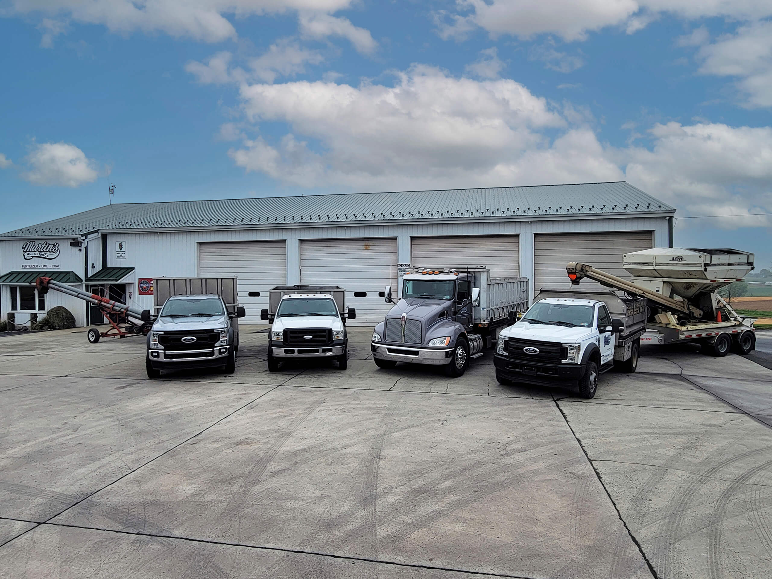 5.16.26 Country Meadows Coal Gallery 1 Four trucks are parked side by side on a concrete lot in front of a large building with four garage doors under a partly cloudy sky.