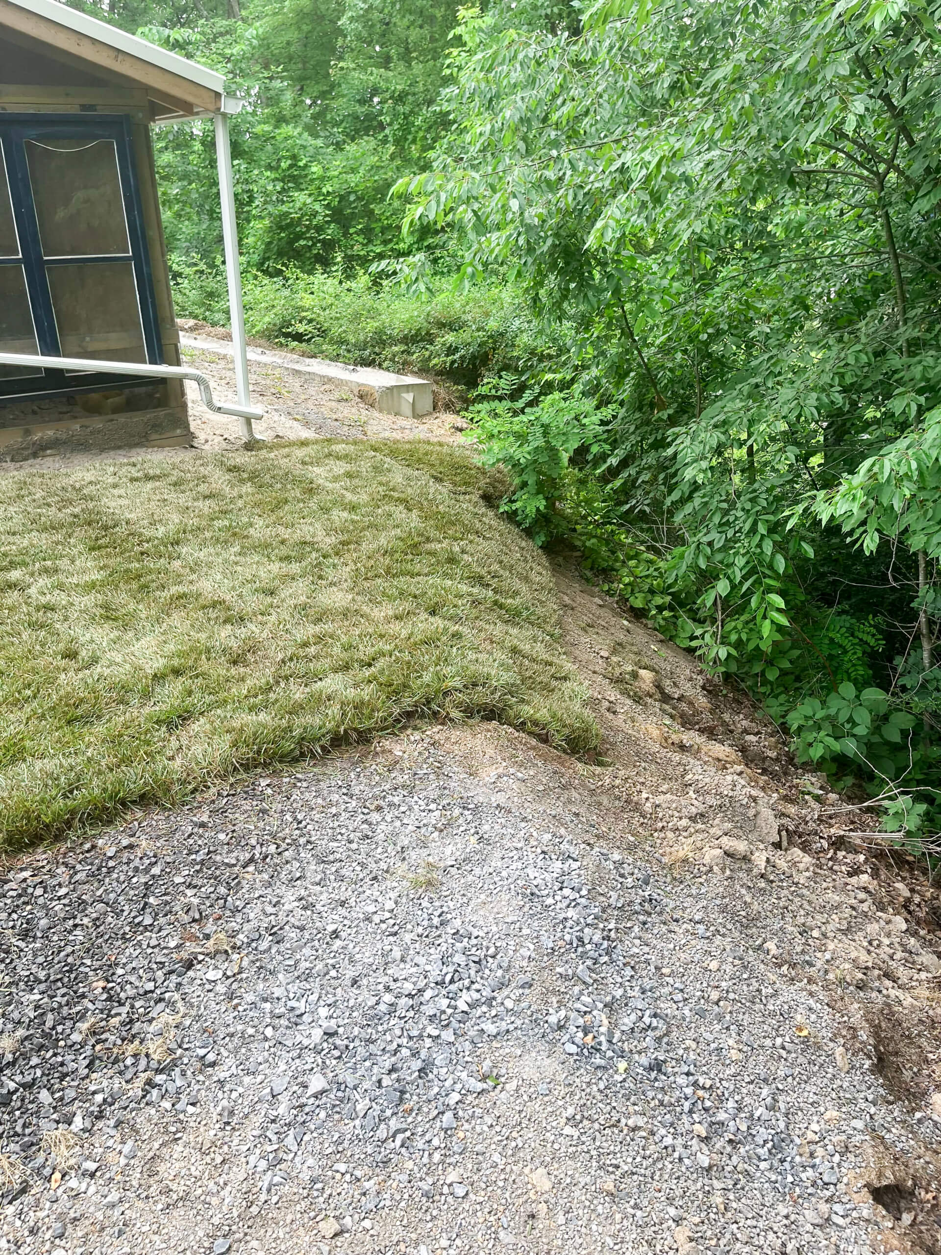 Town's Edge Garden Center New Holland, PA A gravel path meets a patch of fresh sod next to a house, with a sloped dirt area leading down to dense green woods.