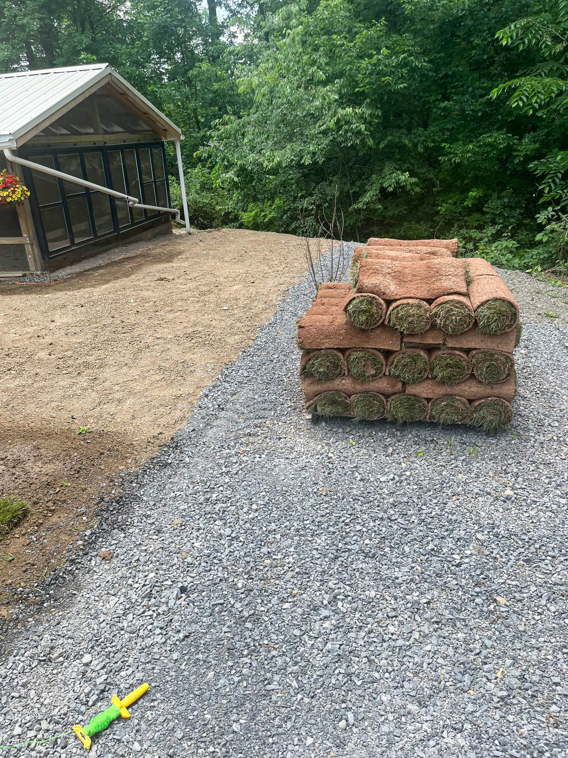 Town's Edge Garden Center New Holland, PA Rolls of sod stacked on gravel near a greenhouse, with a patch of bare dirt beside them and a child's toy shovel on the ground. Trees and greenery are in the background.