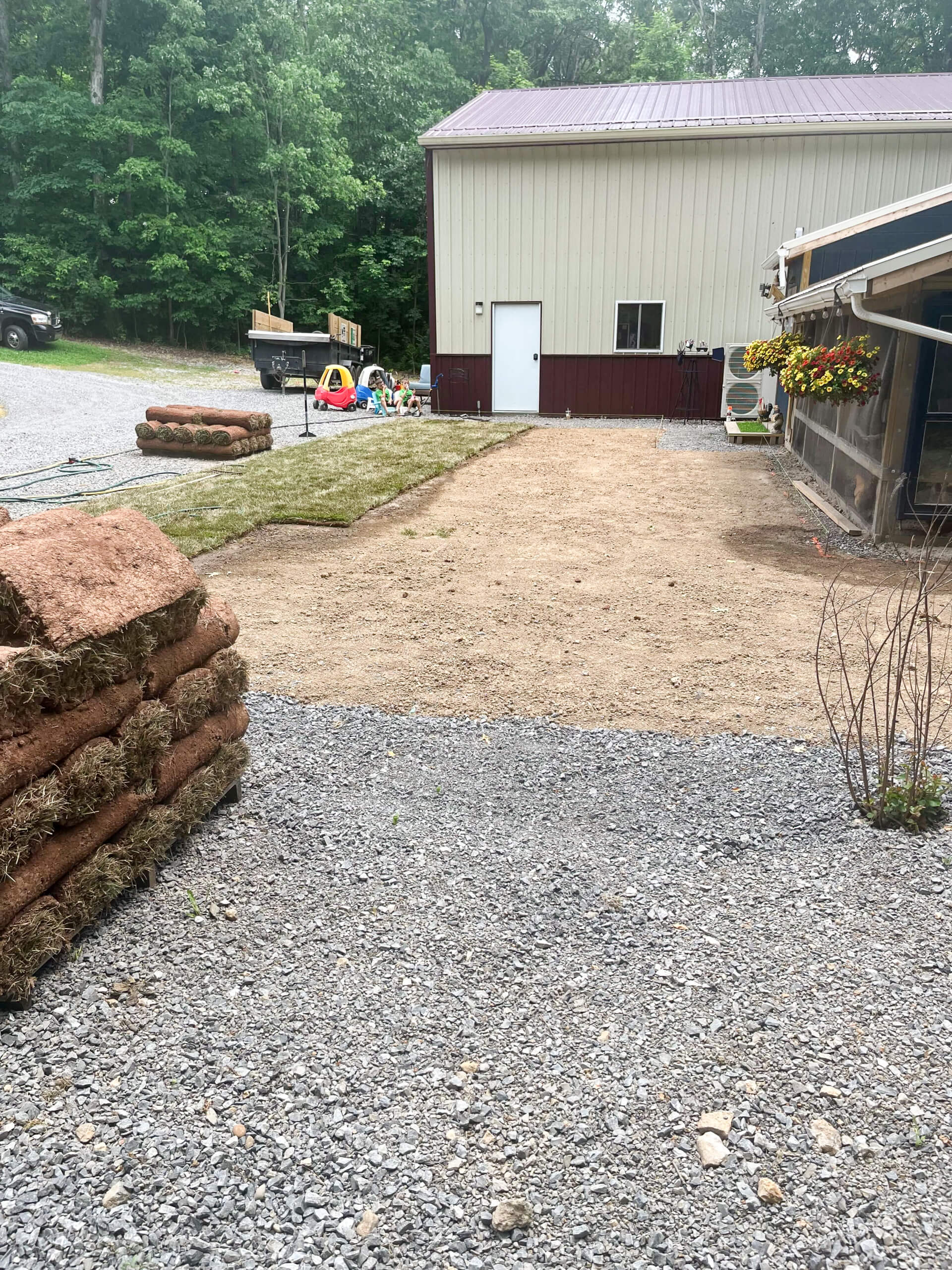 Town's Edge Garden Center New Holland, PA Stacks of sod are on gravel near a patch of bare soil beside a metal building; children's toys and lawn equipment are seen in the background.