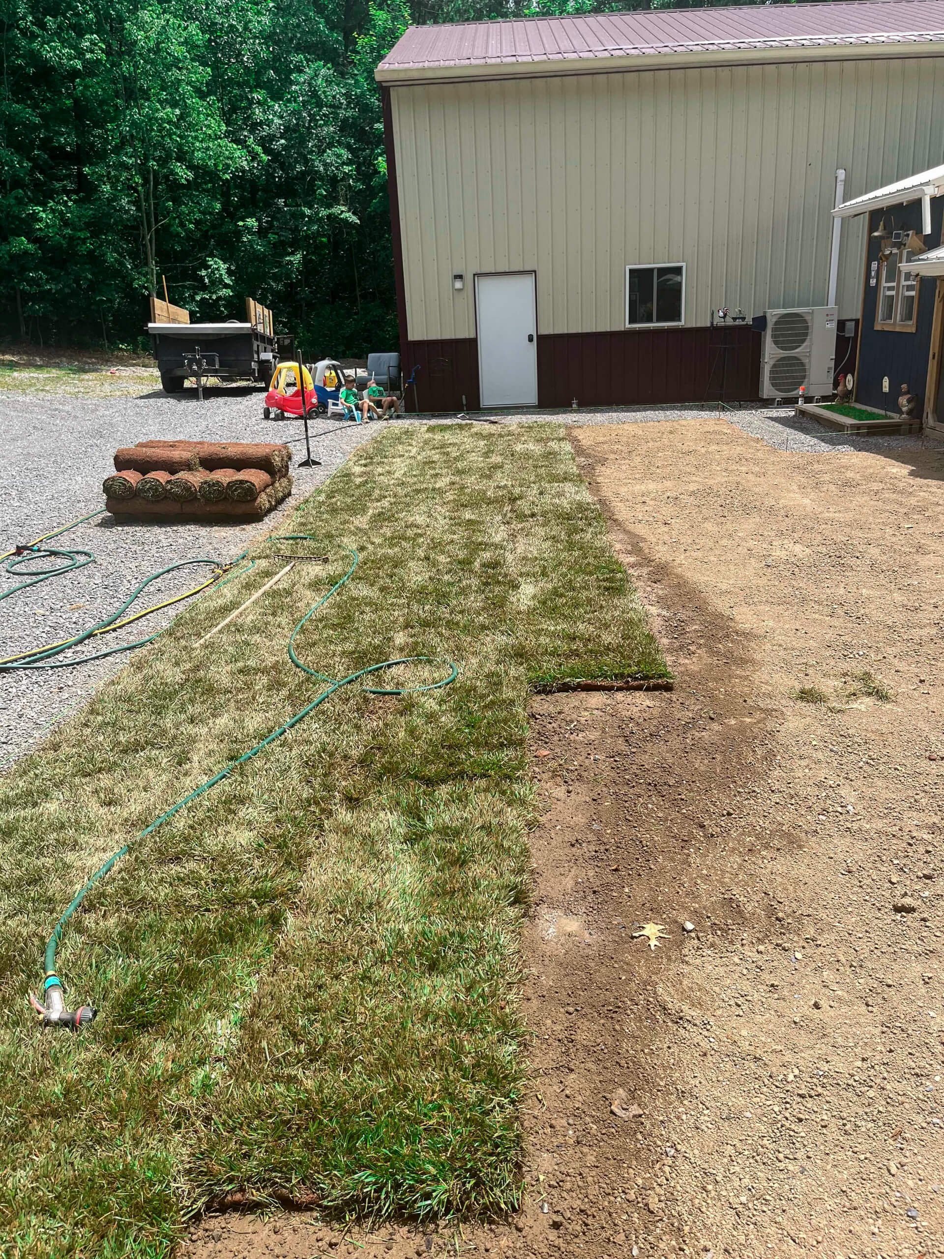 Town's Edge Garden Center New Holland, PA Fresh sod is being laid next to a building; a hose and sprinkler are watering part of the newly installed grass, with more sod rolls and tools visible nearby.