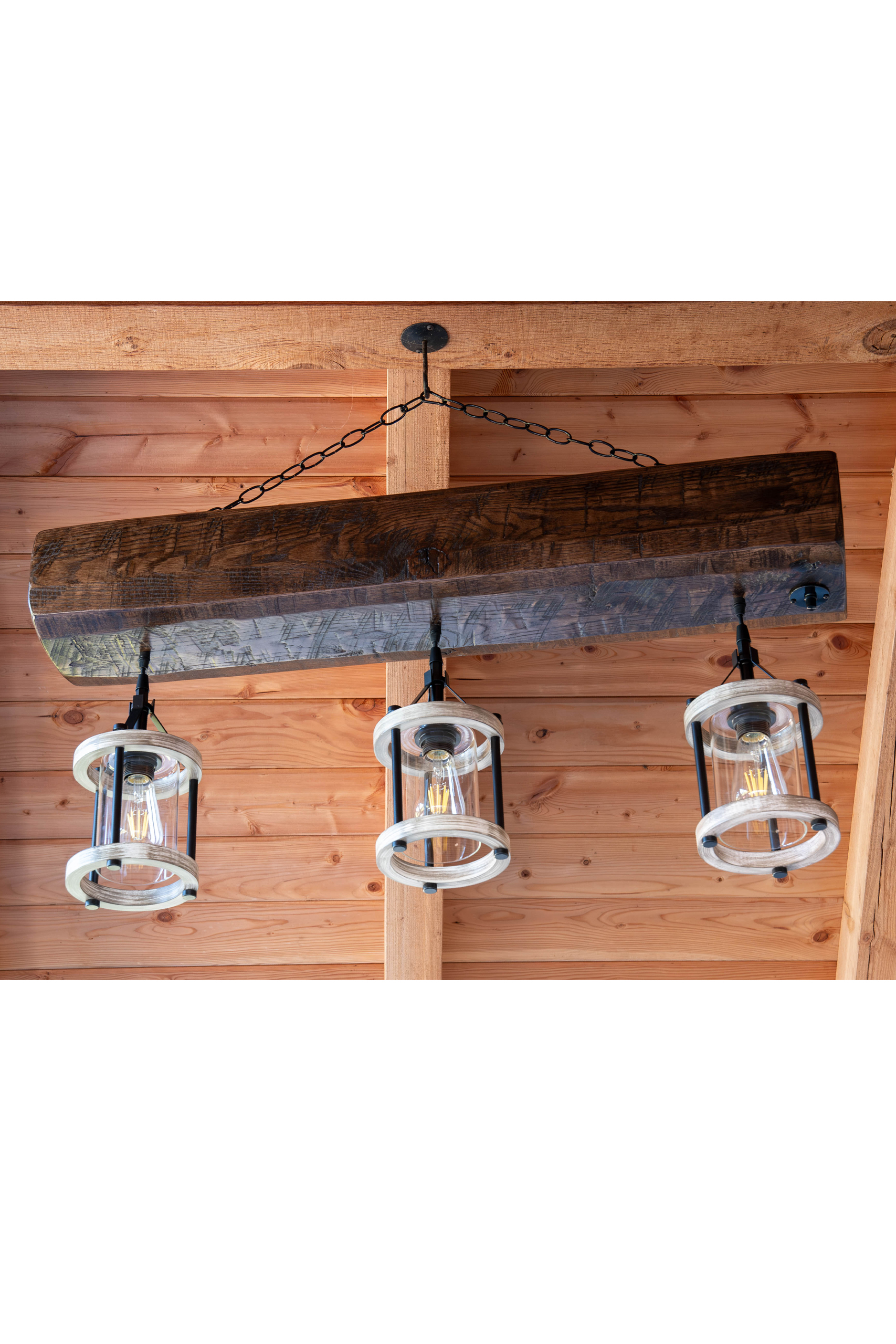 7.8.26 Spring Valley Woodworking Light Fixtures Gallery 2 A rustic wooden beam chandelier with three glass lantern-style lights is suspended from a wooden ceiling with a black chain.