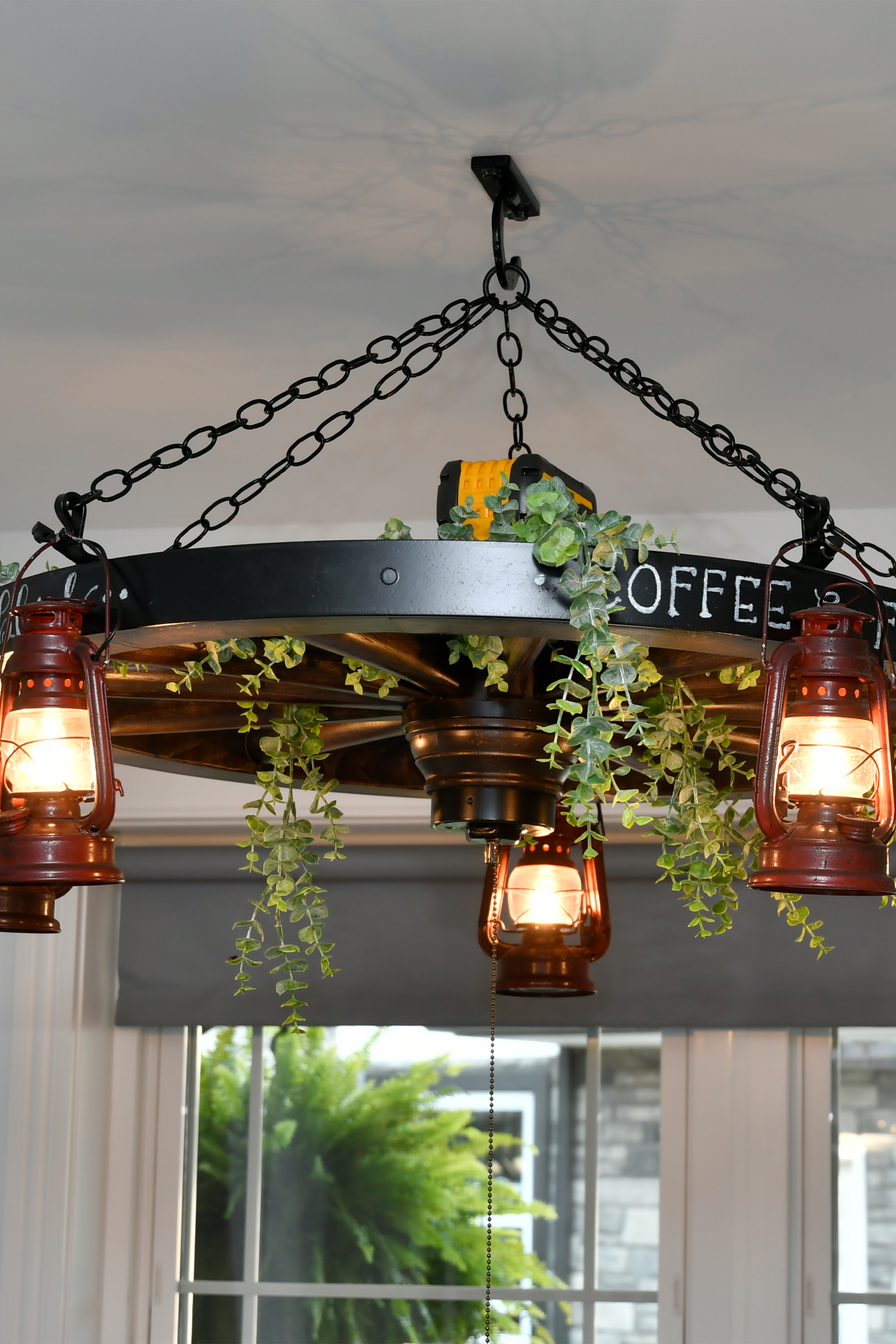 7.8.26 Spring Valley Woodworking Light Fixtures Gallery 3 A chandelier made from a wagon wheel with hanging lantern-style lights, green foliage, and a "COFFEE" sign, suspended by chains from the ceiling.