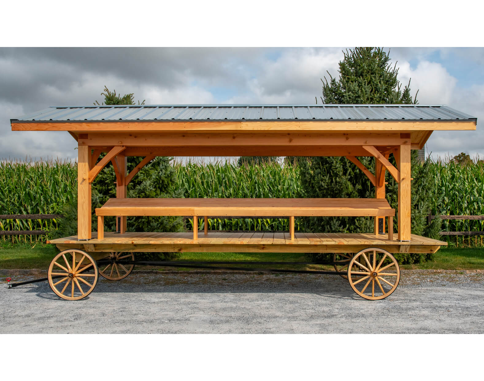 7.8.26 Spring Valley Woodworking Wagons Gallery 3 A wooden wagon with a metal roof and benches is parked on gravel beside a cornfield, with trees and a cloudy sky in the background.
