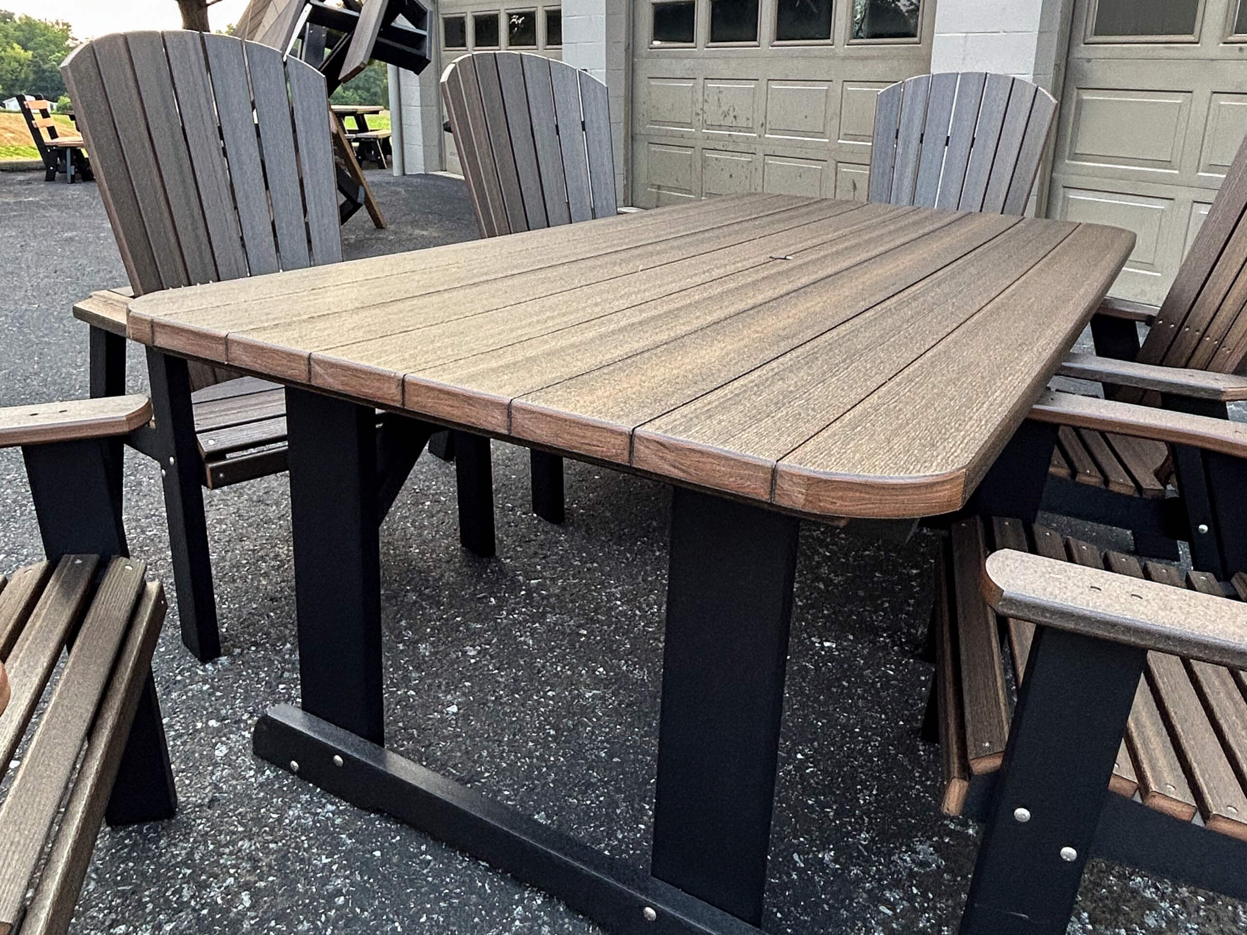 Oregan Lawn Furniture Leola, Pa A rectangular outdoor table with a wood-like surface and black legs, surrounded by four matching chairs, sits on an asphalt surface near a building with garage doors.
