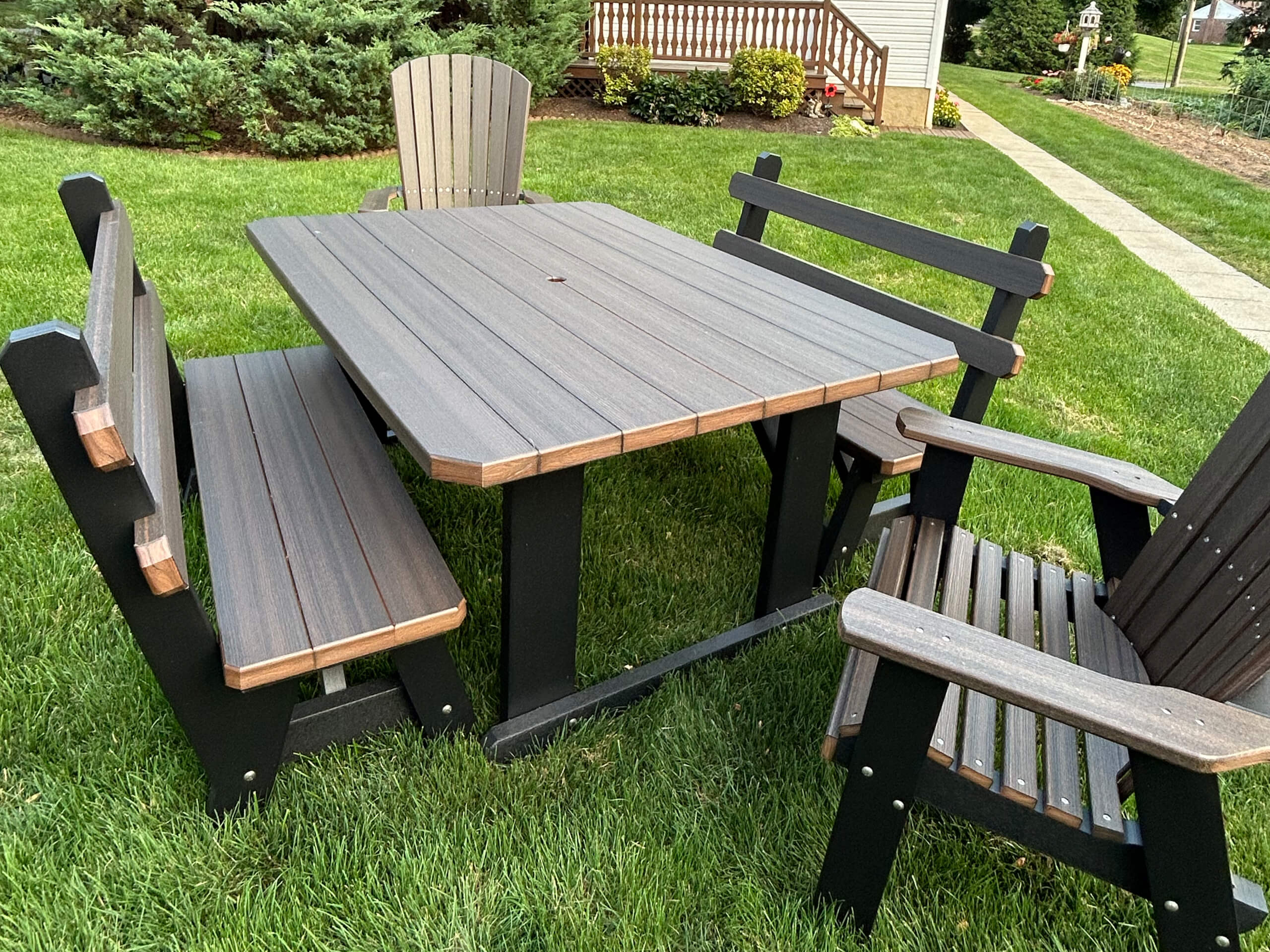Oregan Lawn Furniture Leola, Pa A wooden outdoor table with two benches and two armchairs is placed on a green lawn near a house and garden walkway.