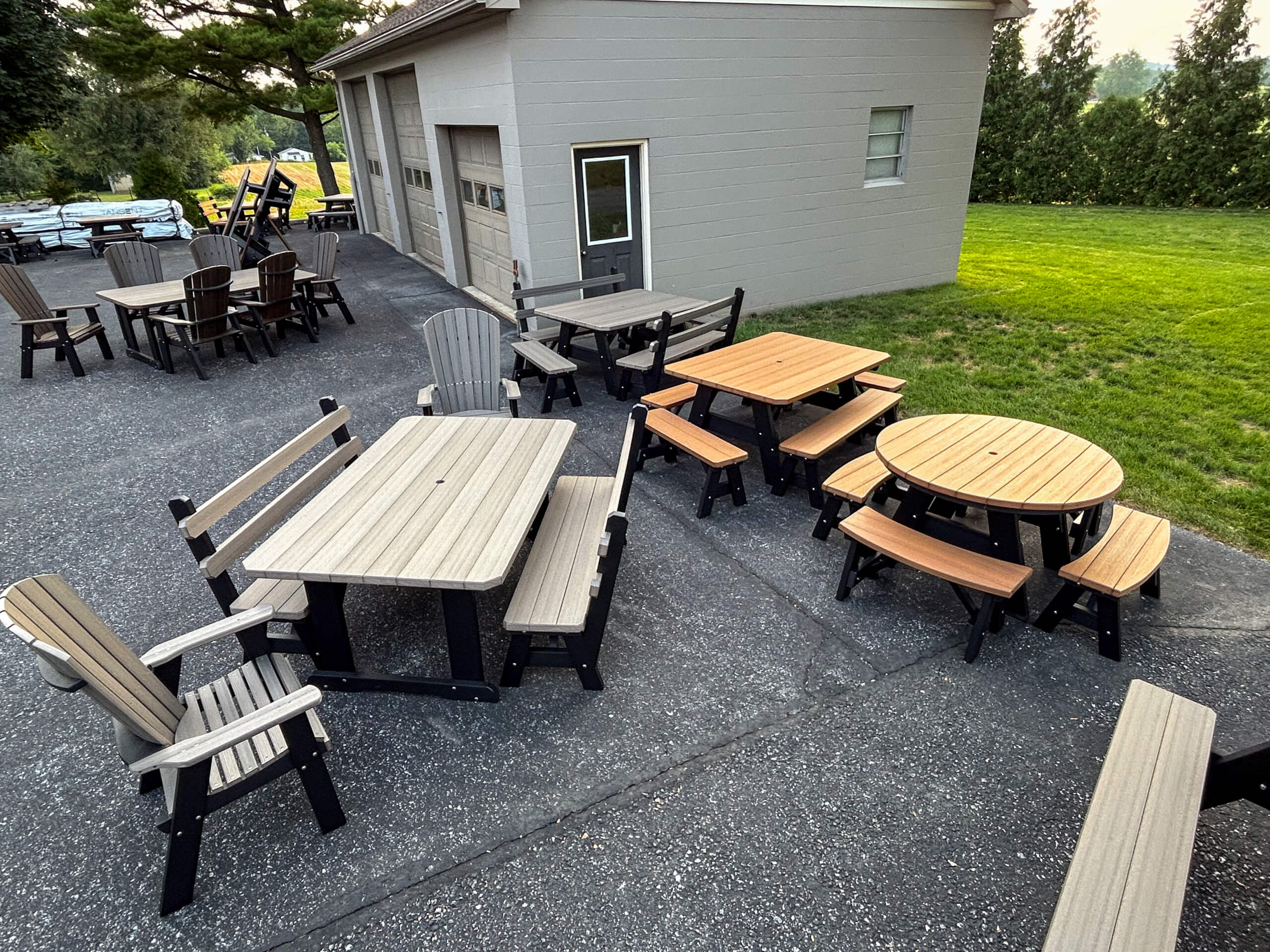 Oregan Lawn Furniture Leola, Pa Outdoor patio area with several wooden tables and chairs, including rectangular and round tables with benches, set on a paved surface near a gray building and grassy area.
