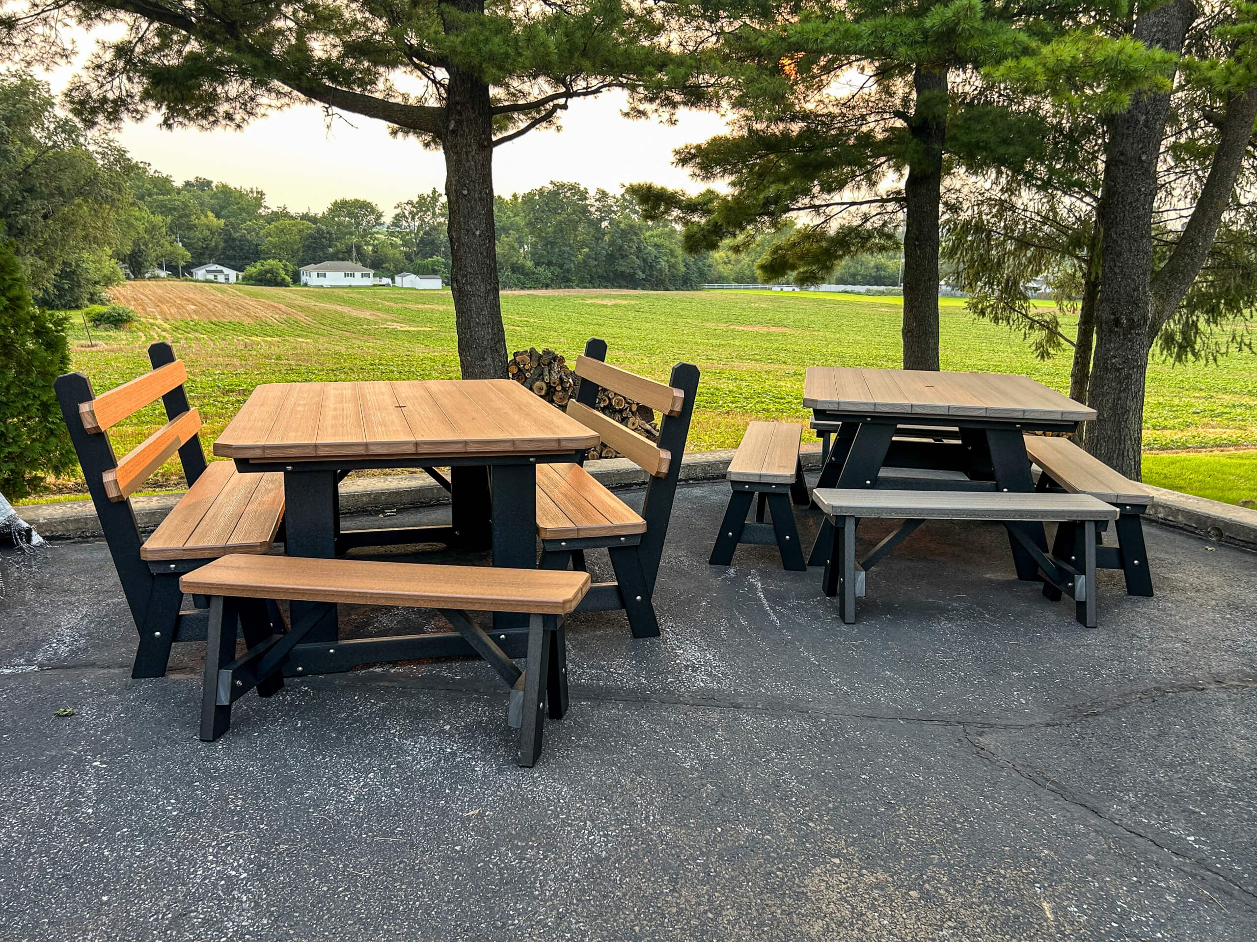 Oregan Lawn Furniture Leola, Pa Two wooden picnic tables with benches are set on a paved area under trees, overlooking a grassy field and distant houses.