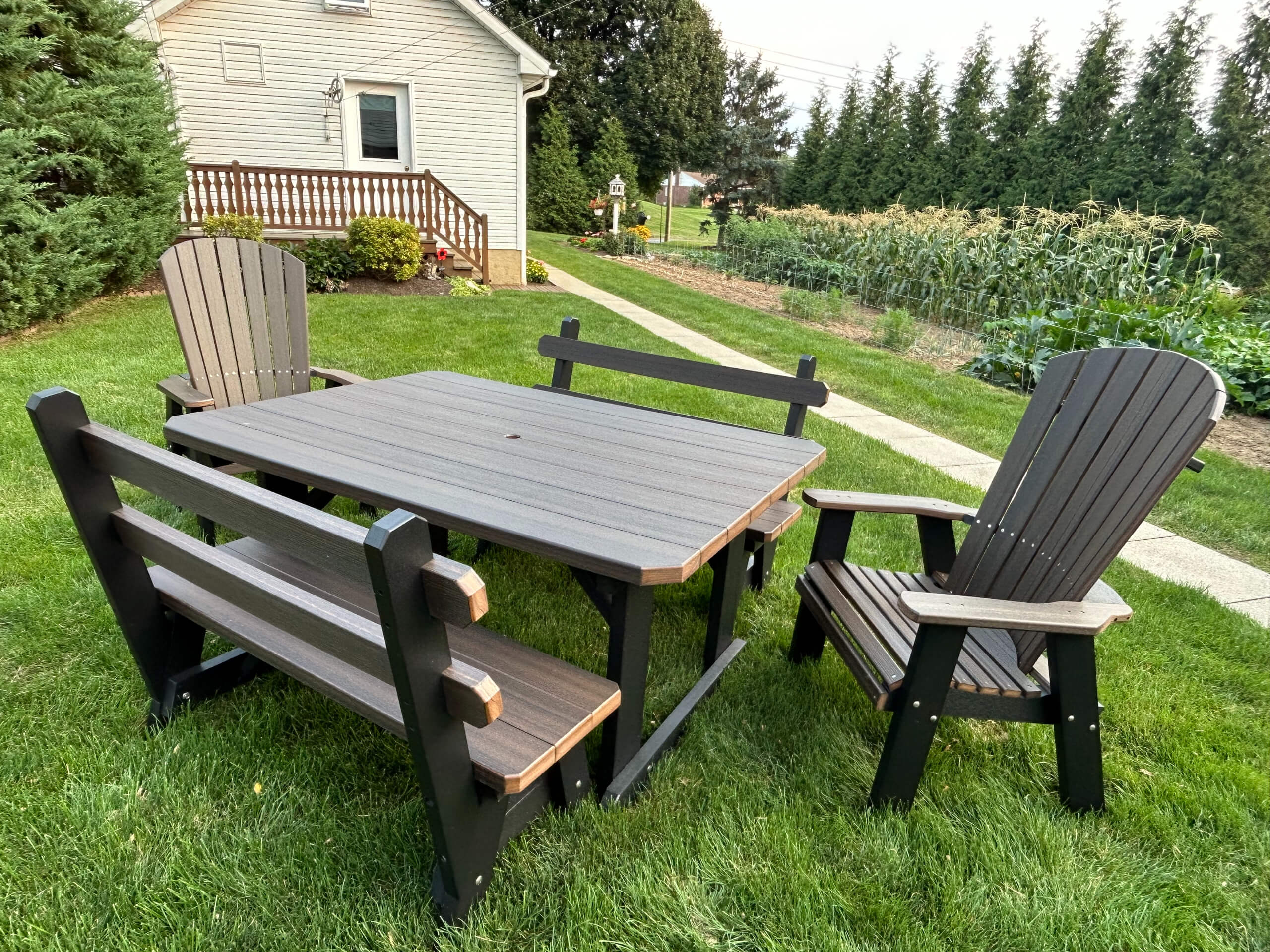 Oregan Lawn Furniture Leola, Pa Outdoor patio set with a rectangular table, two Adirondack chairs, and two benches on a grassy lawn near a house and a vegetable garden.