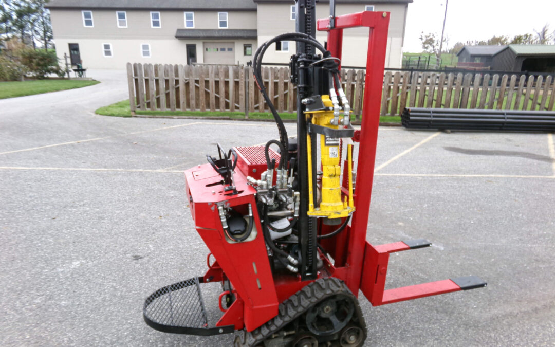 9.9.26 Nickel Mine Welding Gallery 1-12 Red, tracked hydraulic machine with metal forks and yellow hydraulic arm, parked on pavement in front of a fenced residential area.
