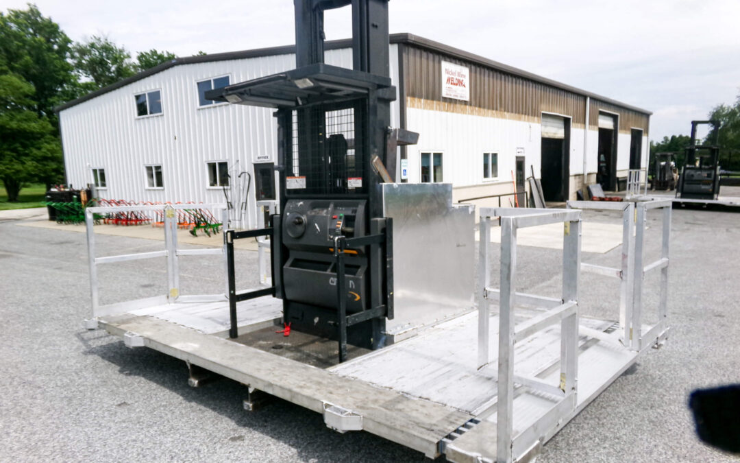 9.9.26 Nickel Mine Welding Gallery 1-6 A forklift is positioned on a large metal loading platform with safety rails, in an outdoor industrial area near white warehouse buildings.